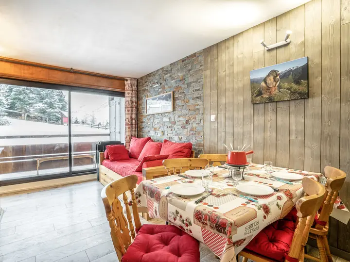 Hyggelig spiseplads med en rød sofa og et spisebord sat til et måltid. Væggene er en blanding af træ og sten. Et billede af et dyr hænger over sofaen. Store glasskydedøre giver udsigt til et snelandskab udenfor.