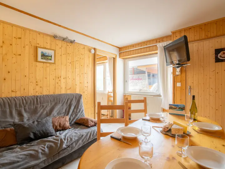 Hyggeligt værelse med trævægge med et rundt spisebord med tallerkener og glas, en flaske vin og ost. En grå sofa med hynder er ved siden af, og et væghængt tv er synligt. Naturligt lys strømmer gennem to vinduer.