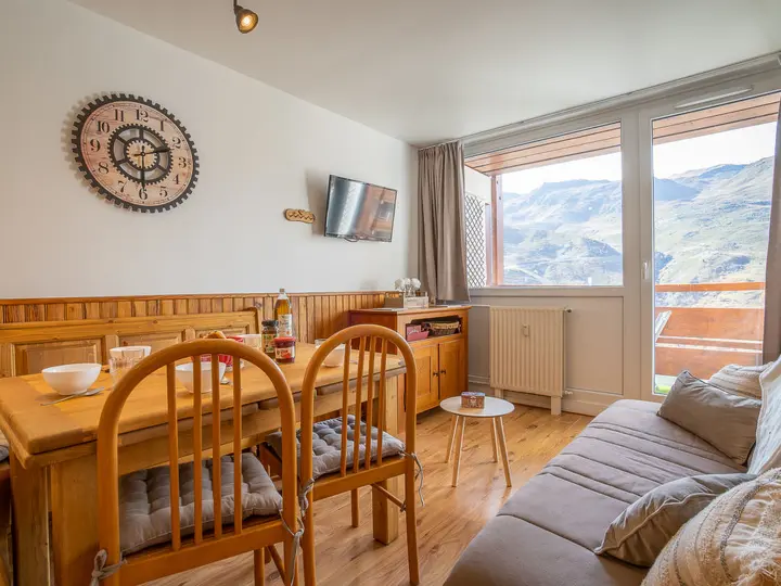 Hyggelig spiseplads med træmøbler, med et rundt ur på væggen og et fjernsyn. Store vinduer tilbyder en naturskøn udsigt over bjergene. Et lille bord med en flaske og kopper og en behagelig sofa med puder fuldender rummet.