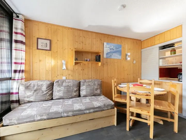 Hyggeligt opholdsrum med trævægge, med en gråmønstret sofa, et rundt spisebord med stole og vægkunst. Et delvist synligt køkken er i baggrunden. Naturligt lys kommer ind gennem et vindue med plaidgardiner.