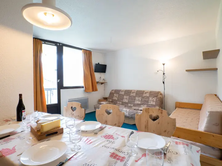 En hyggelig opholds- og spisestue med et spisebord i træ til fire. Værelset har en sofa, en daybed og et lille væghængt tv. Orange gardiner komplementerer de hvide vægge. En loftlampe og gulvlampe giver belysning.