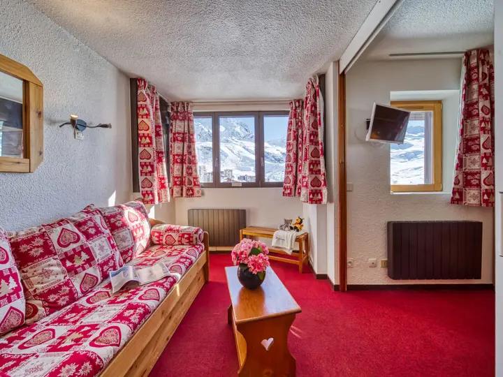 Hyggelig alpestue med en rød og hvid hjertemønstret sofa, rød løber og blomstergardiner. Et sofabord af træ rummer en vase med blomster. Stort vindue afslører snedækkede bjerge udenfor. Varm belysning bidrager til den indbydende atmosfære.