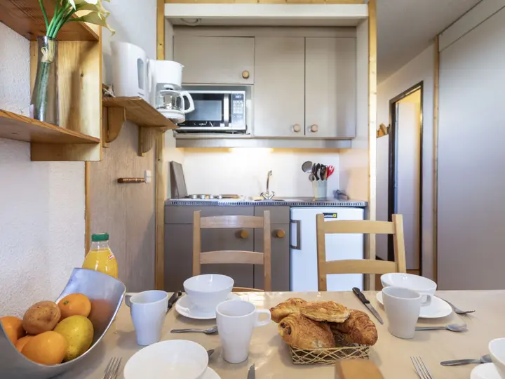 Et lille køkken med lyse træskabe, mikroovn, kaffemaskine og vask. Et bord er dækket til fire med hvide fade, kopper, croissanter, frugt og appelsinjuice. En vase med blomster står på en hylde.