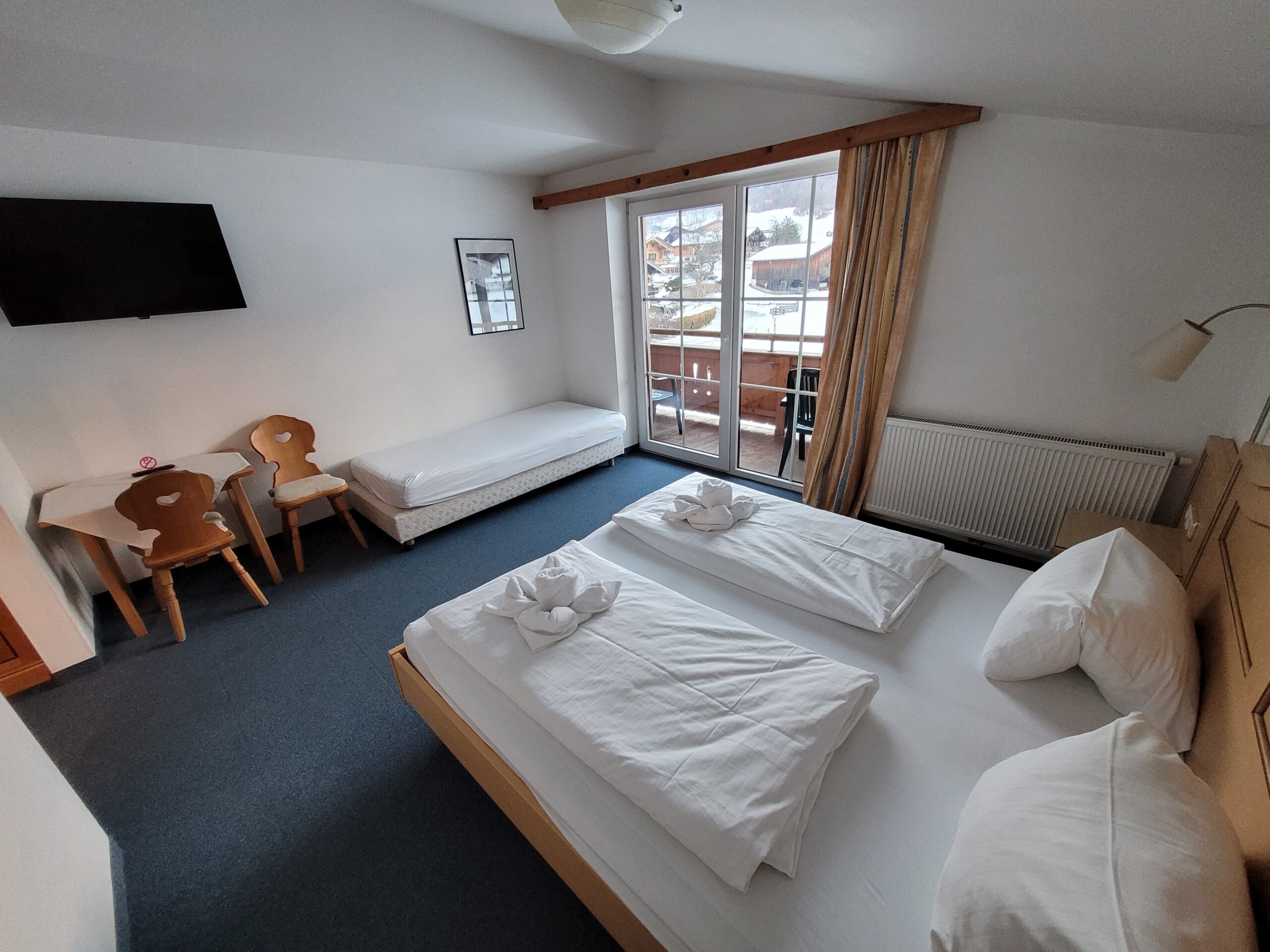 Et pænt hotelværelse med en dobbeltseng, en enkeltseng, et vægmonteret tv, to træstole ved et lille bord og glasdøre, der fører ud til en balkon med udsigt over sneklædte bjerge. Håndklæder ligger foldet sammen på sengene.