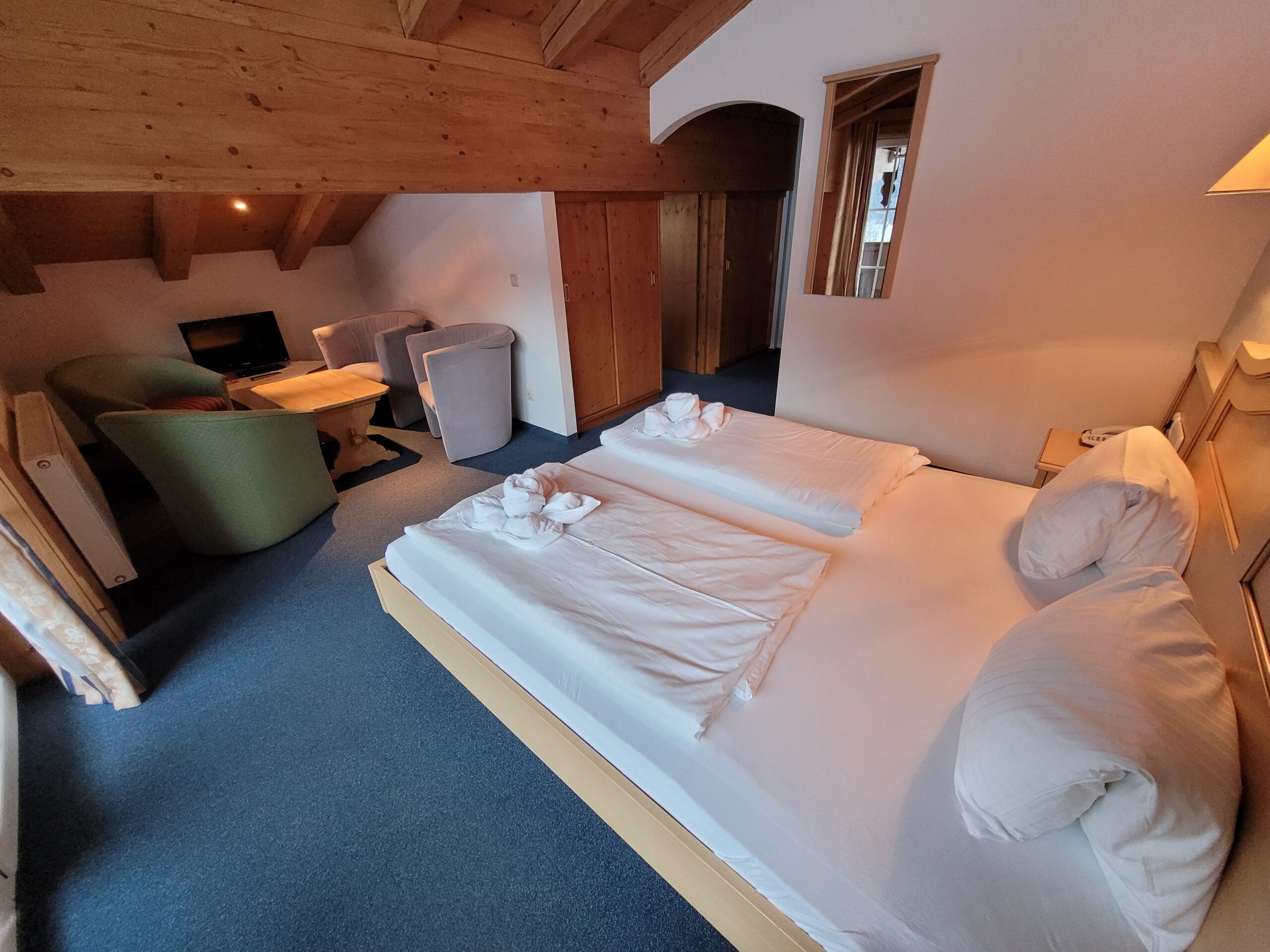 Hyggeligt hotelværelse med dobbeltseng, to lænestole omkring et lille bord, et tv og træbjælker i loftet. Håndklæder er foldet sammen på sengen, og naturligt lys kommer ind gennem et vindue.