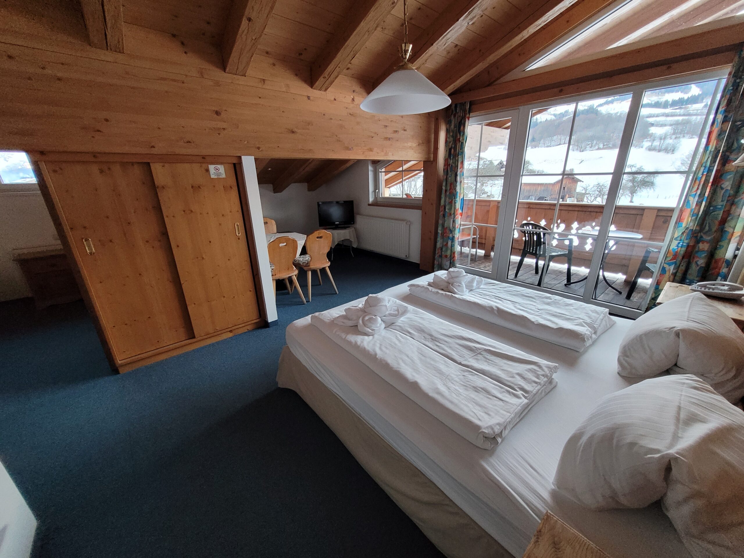 Et hyggeligt hotelværelse med træloft og -møbler, to enkeltsenge ved siden af hinanden, et tv, to stole, en balkon med udsigt over sneen og sammenfoldede håndklæder på sengene.