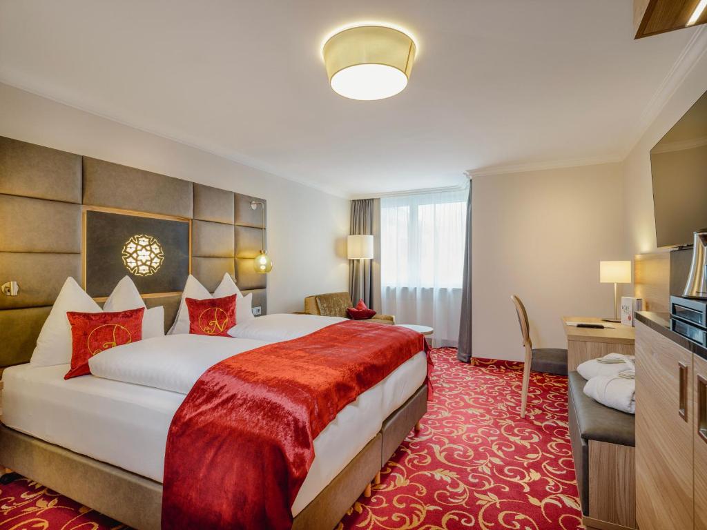 Et hotelværelse med to senge, rødt og hvidt sengetøj, dekorative puder, et mønstret rødt tæppe, et vindue med gardiner, et skrivebord med en stol, en lampe og et fladskærms-tv på et træskab.