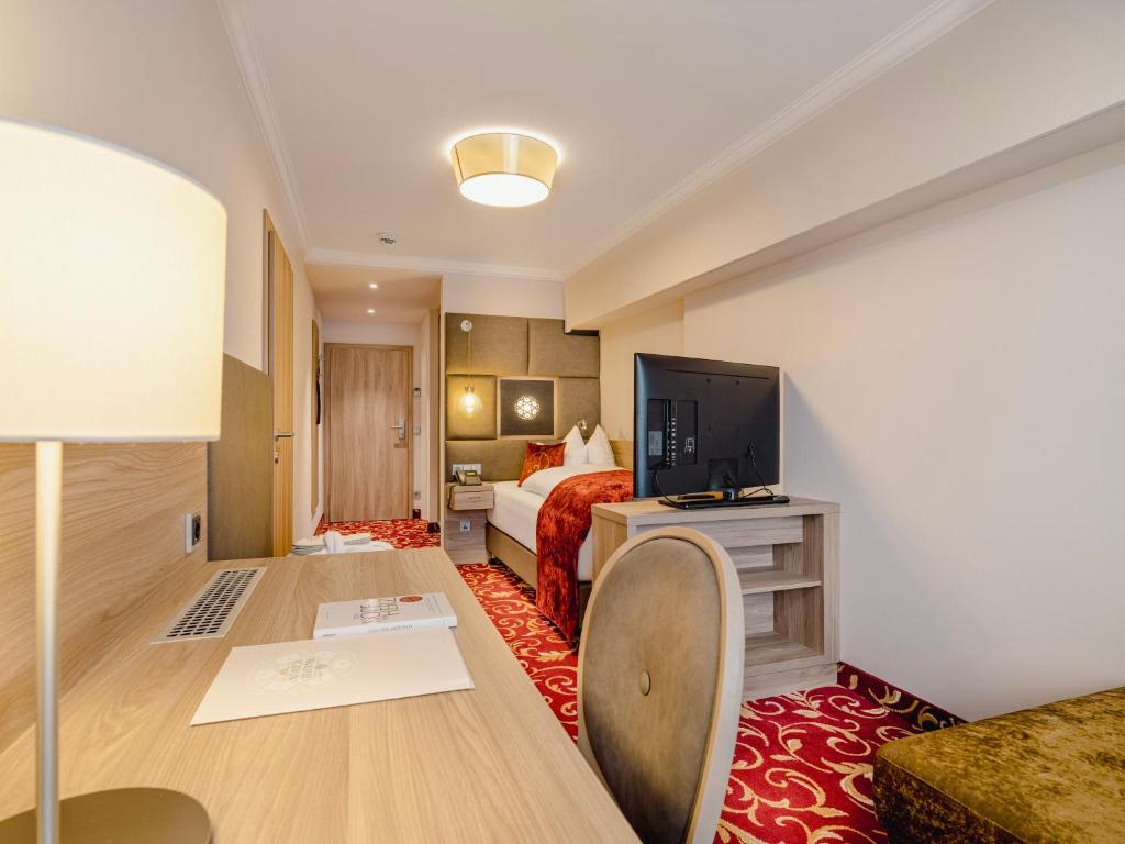 Et moderne hotelværelse med et rødt mønstret tæppe, en seng med hvidt sengetøj og rødt tæppe, et træskrivebord med en stol, en lampe, et tv på et stativ og neutrale vægge.