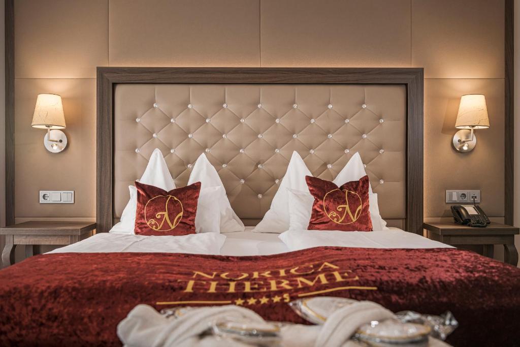 En pænt redt hotelseng med en tuftet beige hovedgærde, to røde pyntepuder og et matchende rødt sengetæppe med teksten Norica Therme. To væglamper og natborde med telefon og stikkontakter er placeret på hver side af sengen.