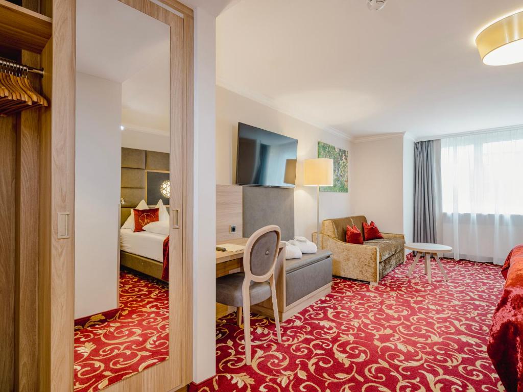 Moderne hotelværelse med et rødt mønstret tæppe, en seng delvist synlig i spejlet, et skrivebord med en stol, en sofa, tv, gulvlampe og et stort vindue med tynde gardiner, der lukker naturligt lys ind.