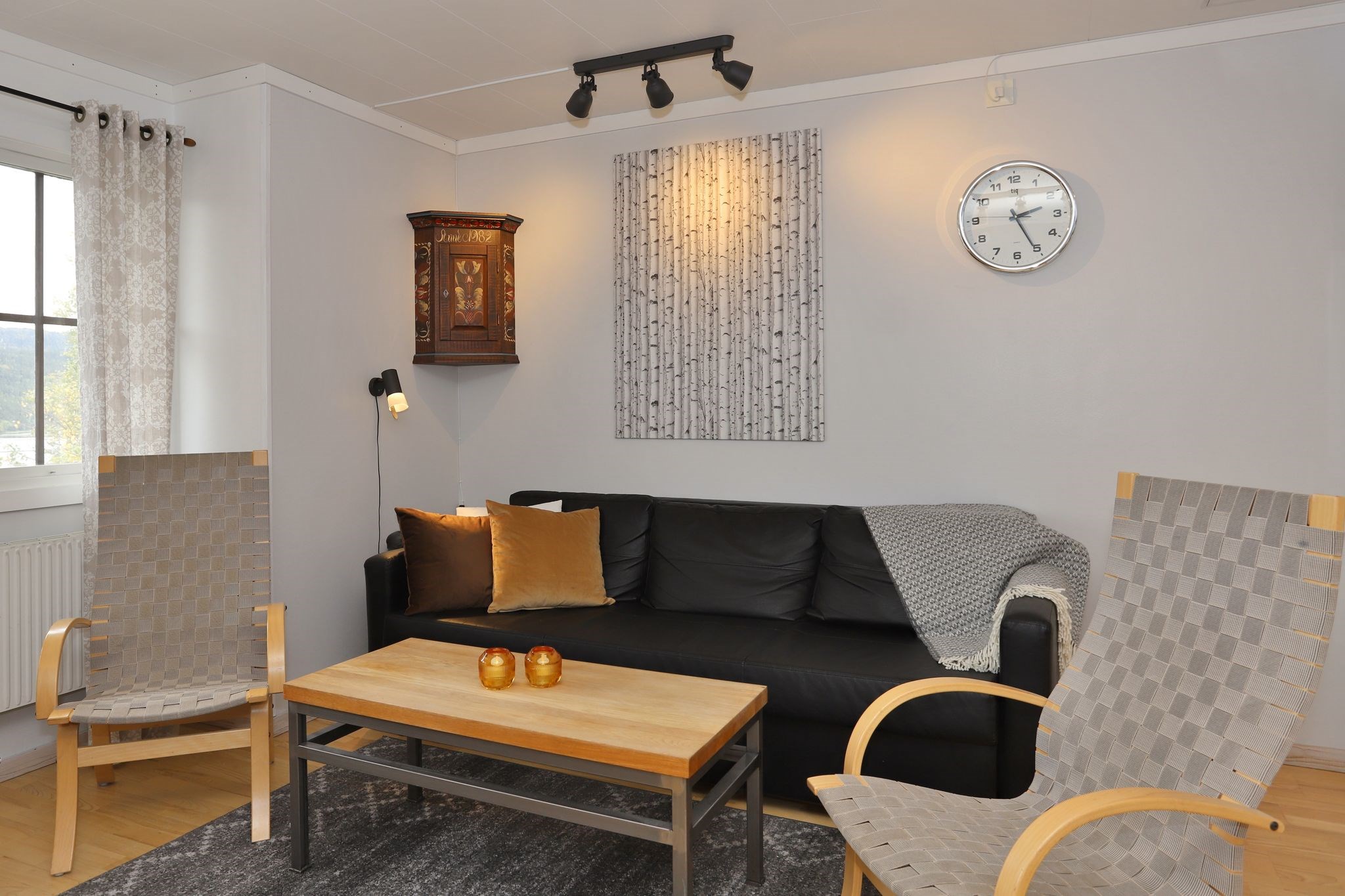 Moderne stue med en sort sofa, to trælænestole, et lyst sofabord i træ, et ur på væggen, en væglampe, et gråt tæppe og dekorativ kunst over sofaen.