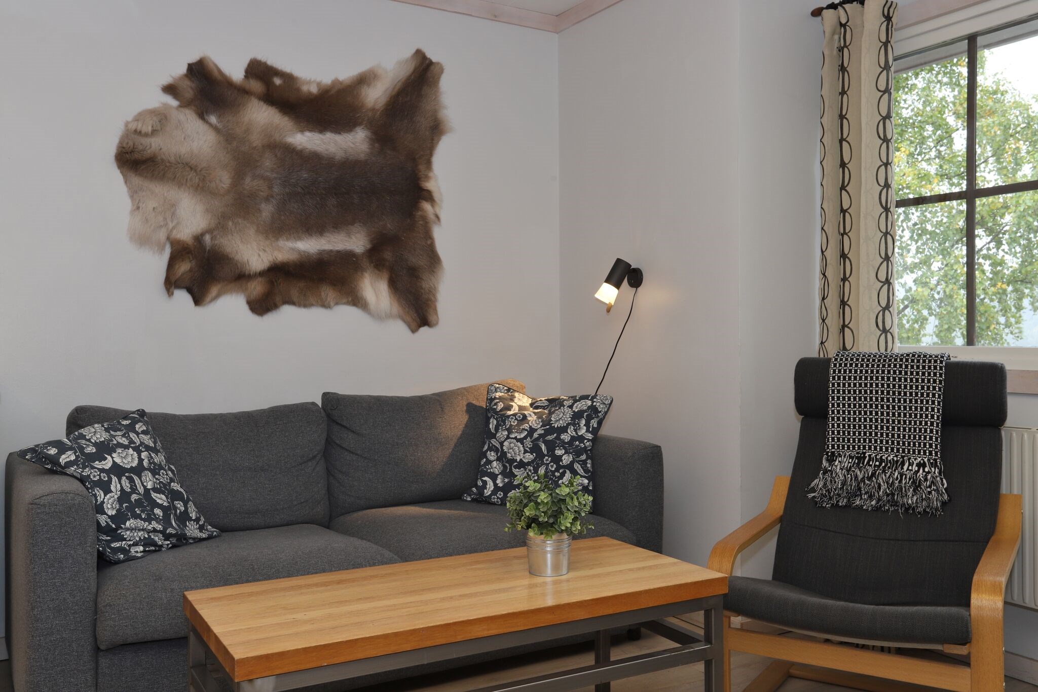 En hyggelig stue med en grå sofa, mønstrede puder, et sofabord i træ med en lille plante, en sort lænestol med et tæppe, en væglampe, et vindue og et pelsmåtte på væggen.