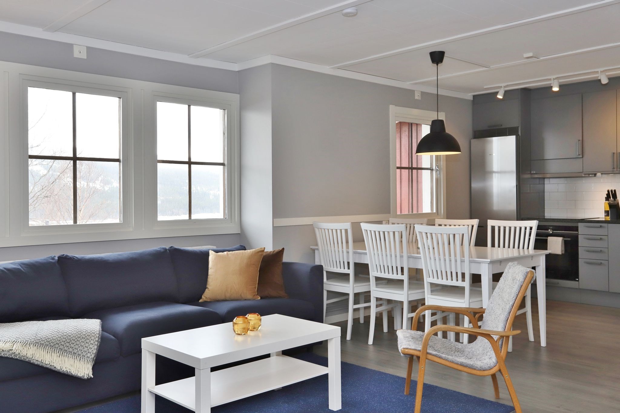 Lys, moderne opholds- og spisestue med grå vægge, blå sofa, hvidt sofabord, træstol, hvidt spisebordssæt og et køkken i baggrunden med grå skabe og apparater i rustfrit stål. Store vinduer lukker naturligt lys ind.