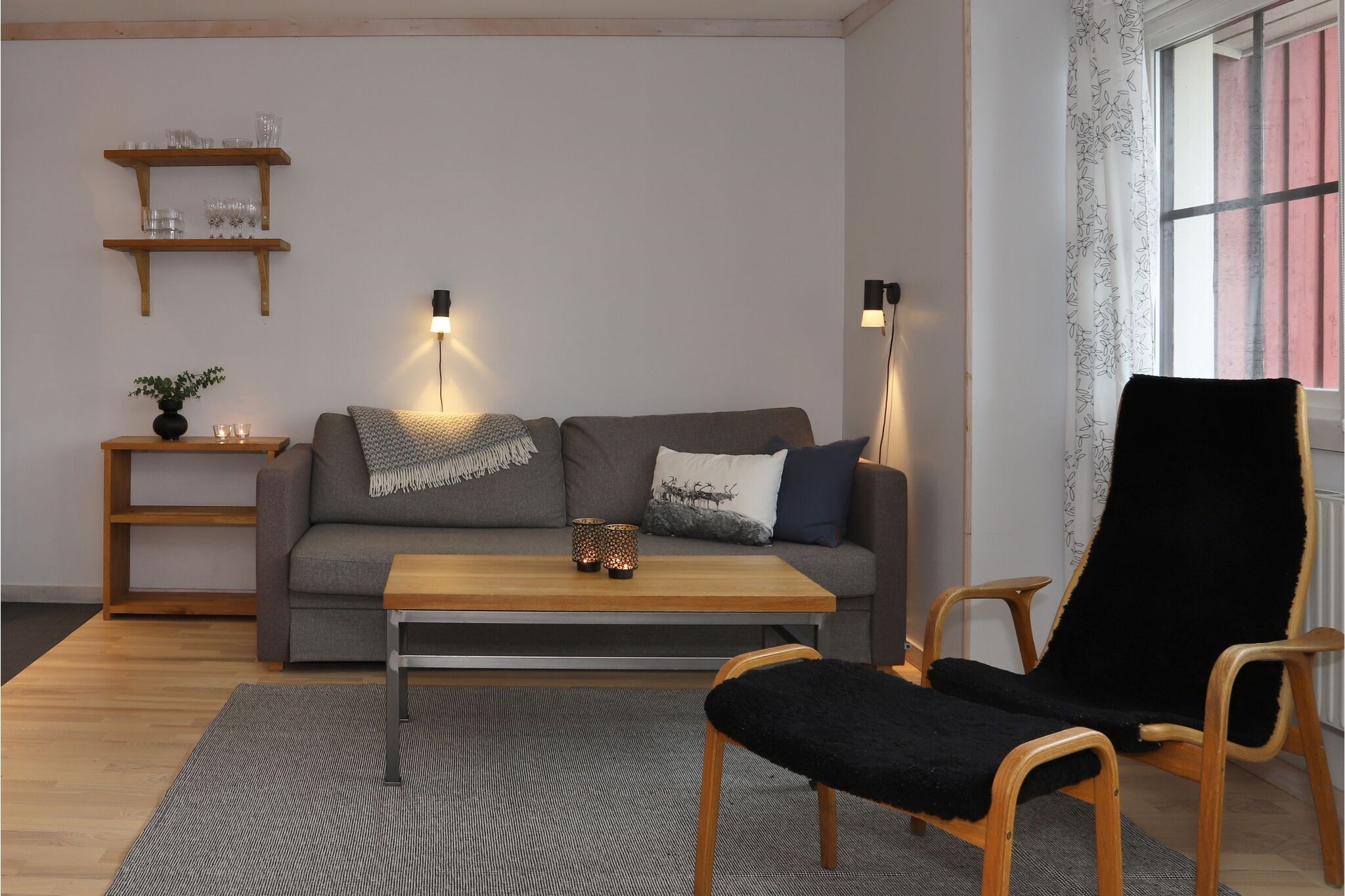 En hyggelig stue med en grå sofa, et sofabord i træ, en sort lænestol, hylder med glas, et lille sidebord med stearinlys og en plante samt et vindue med mønstrede gardiner. Belysningen er varm og indbydende.