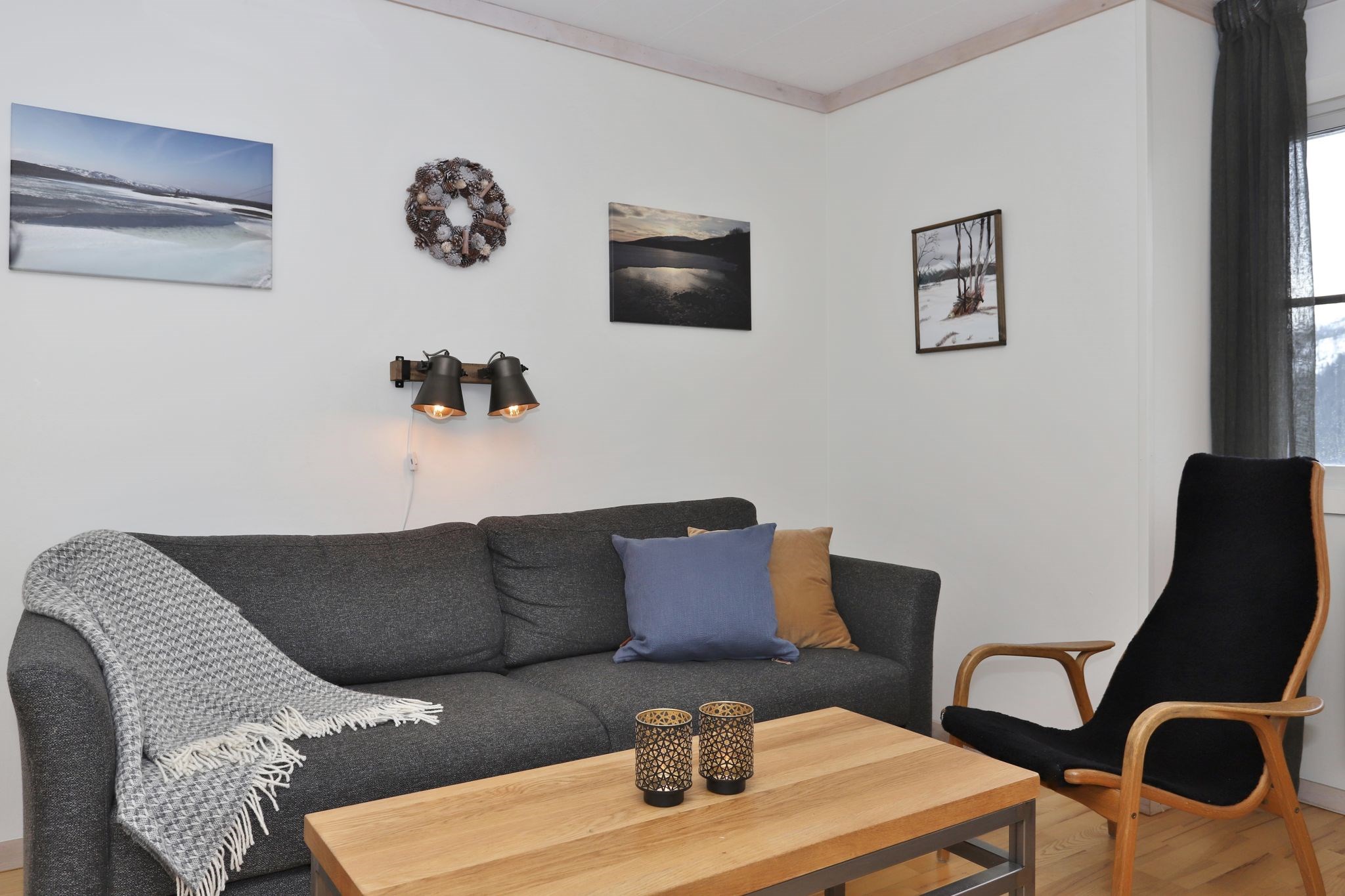 En hyggelig stue med en mørkegrå sofa, blå og beige hynder, et gråt tæppe, en sort lænestol, et sofabord i træ med to lysestager og landskabsfotos på den hvide væg over sofaen.