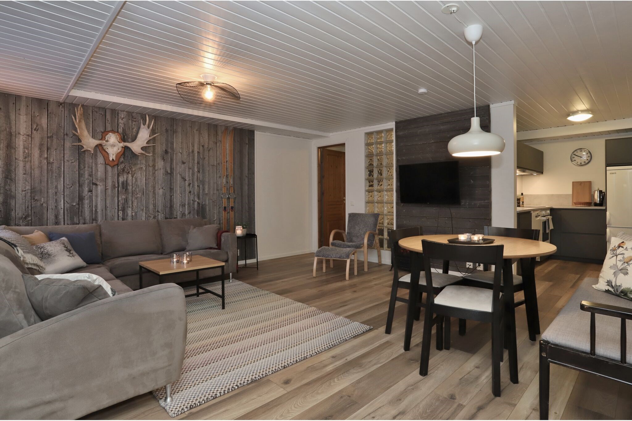 En hyggelig opholds- og spisestue med en grå sofa, trævægge, monterede gevirer, et sort spisebord med stole, et tv og et køkken synligt i baggrunden. Blød belysning og neutral indretning skaber en varm atmosfære.