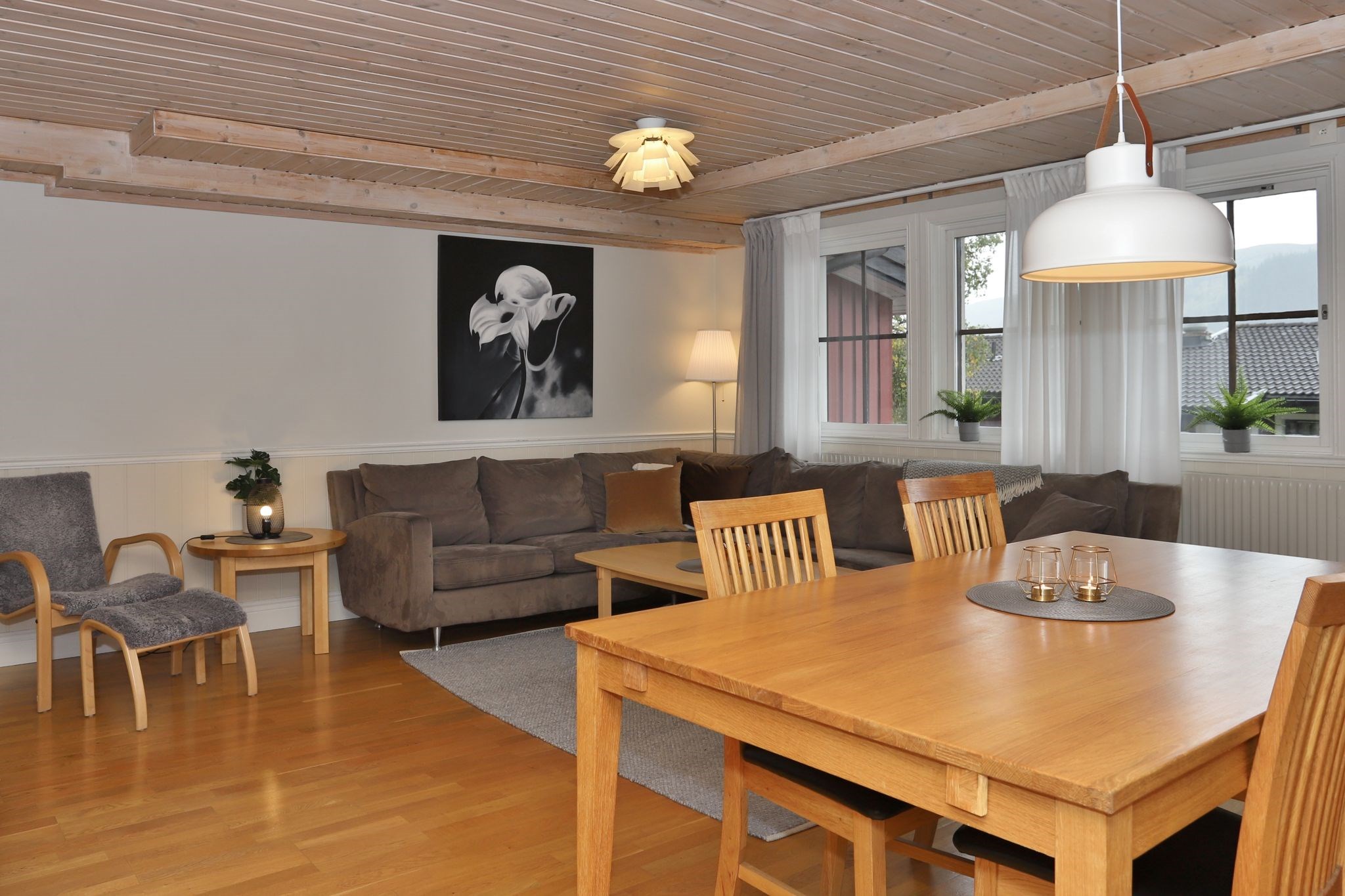En hyggelig opholds- og spisestue med træloft og -gulv, neutralt tonet sofa, lænestole, et spisebord i træ med stole, moderne belysning og et stort vindue med hvide gardiner. Kunstværker pryder væggen over sofaen.