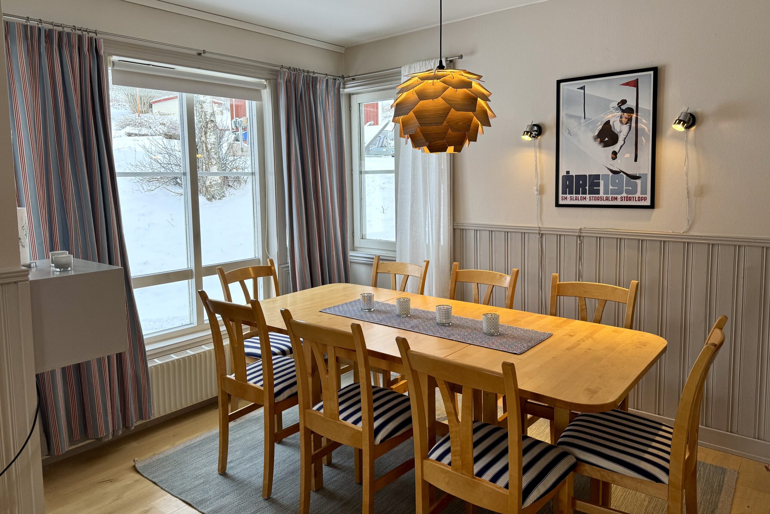 En hyggelig spisestue med et træbord til otte personer, stribede siddehynder, en moderne træpendellampe, store vinduer, der viser sne udenfor, og en vintage skiplakat på væggen.