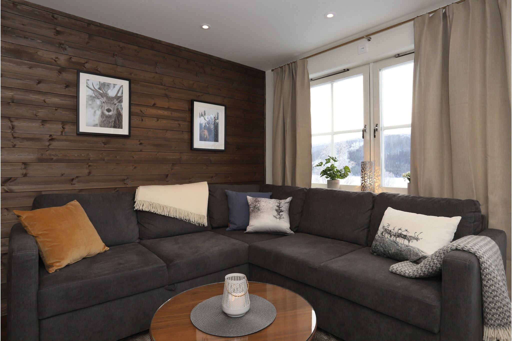 En hyggelig stue med en mørkegrå sofa, diverse puder, et tæppe og et rundt sofabord i træ. Store vinduer med beige gardiner lukker naturligt lys ind. To indrammede billeder hænger på en trævæg.