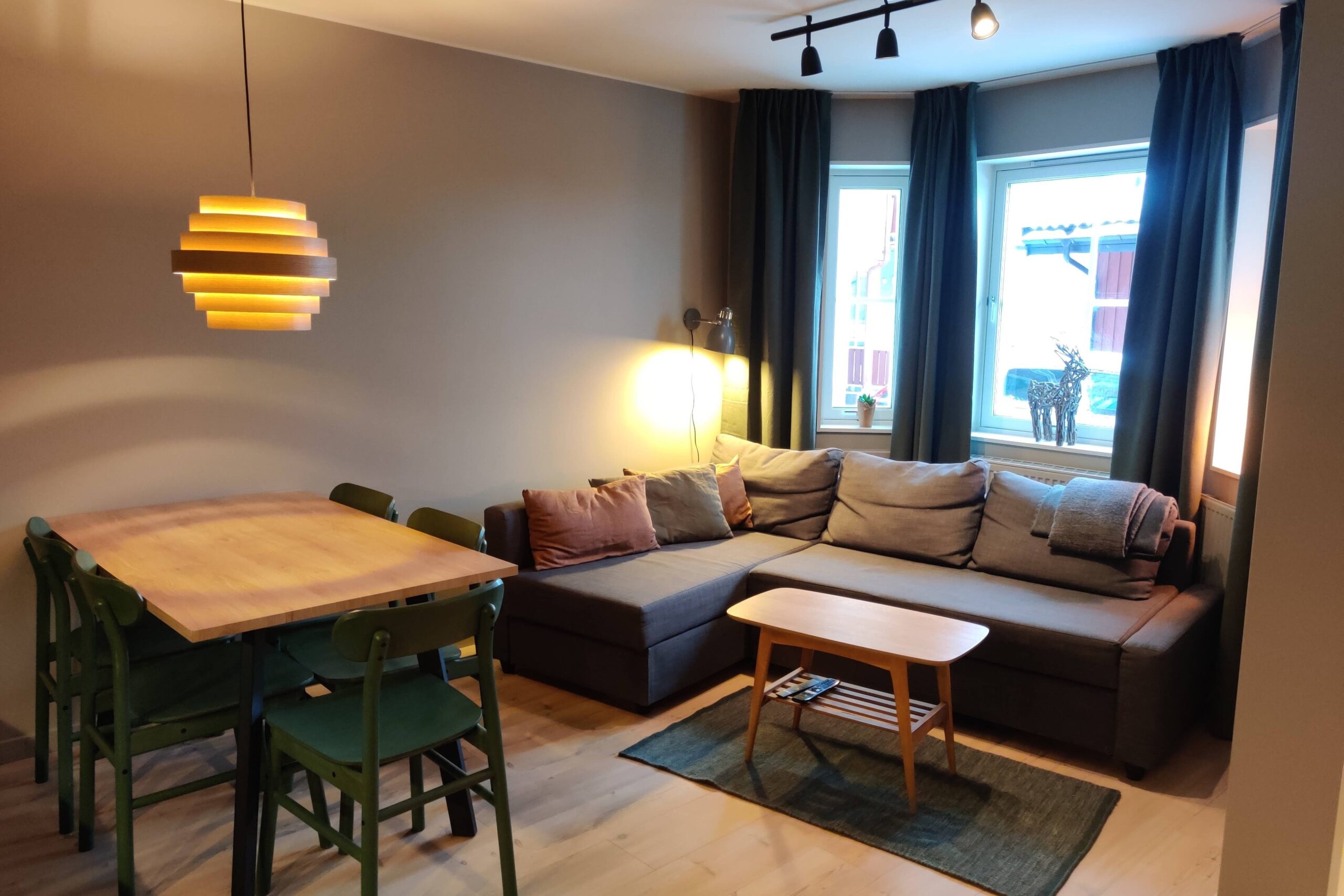 En hyggelig opholds- og spisestue med en grå sofa, et sofabord i træ, grønne stole omkring et spisebord i træ, pendellampe og store vinduer med mørke gardiner, der lukker naturligt lys ind.