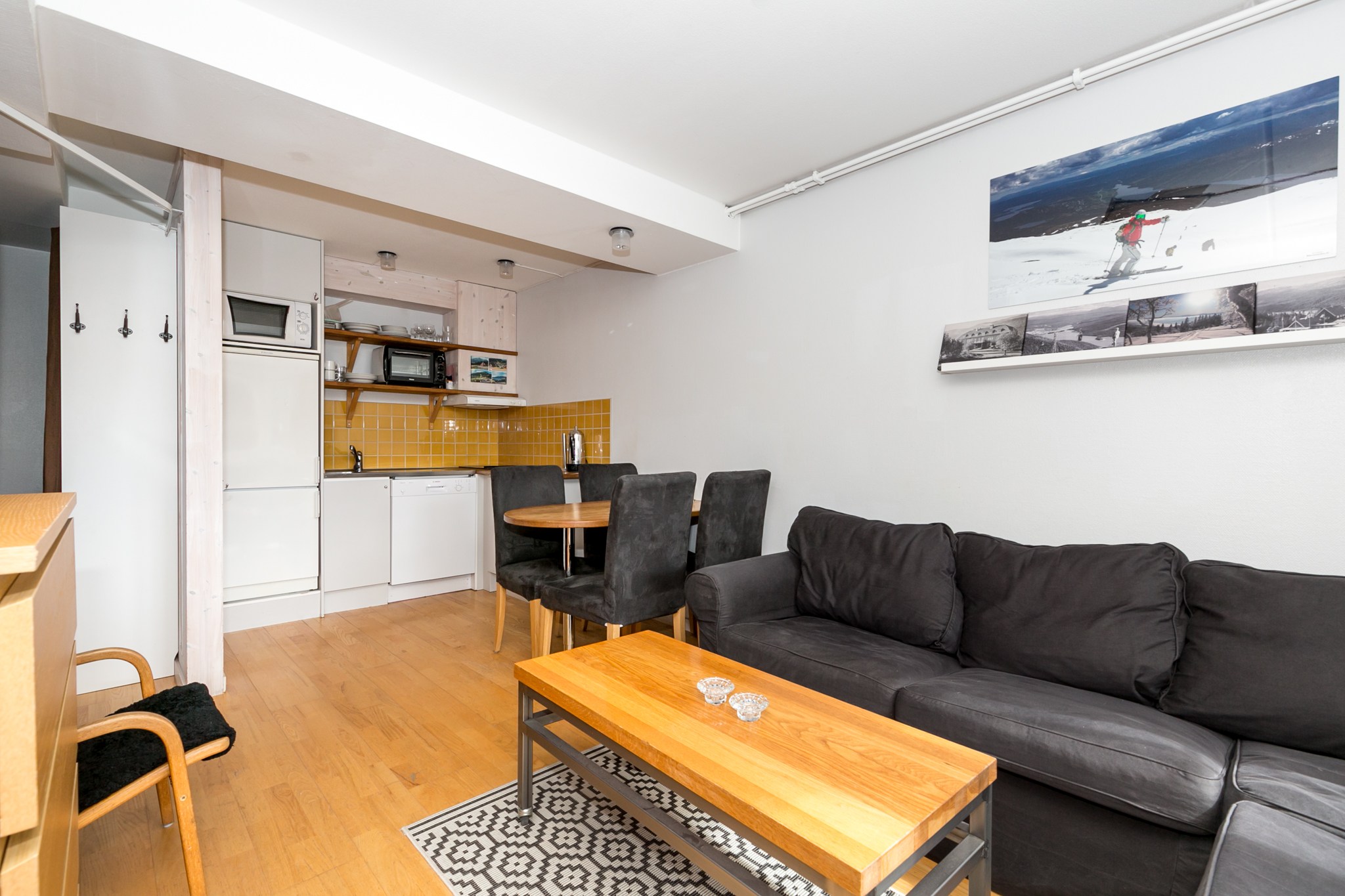 En moderne opholdsstue med en sort sofa, et sofabord i træ og et spisebordssæt. Tekøkkenet har gule fliser, hvide skabe og apparater. Et stort foto af en skiløber pryder væggen.