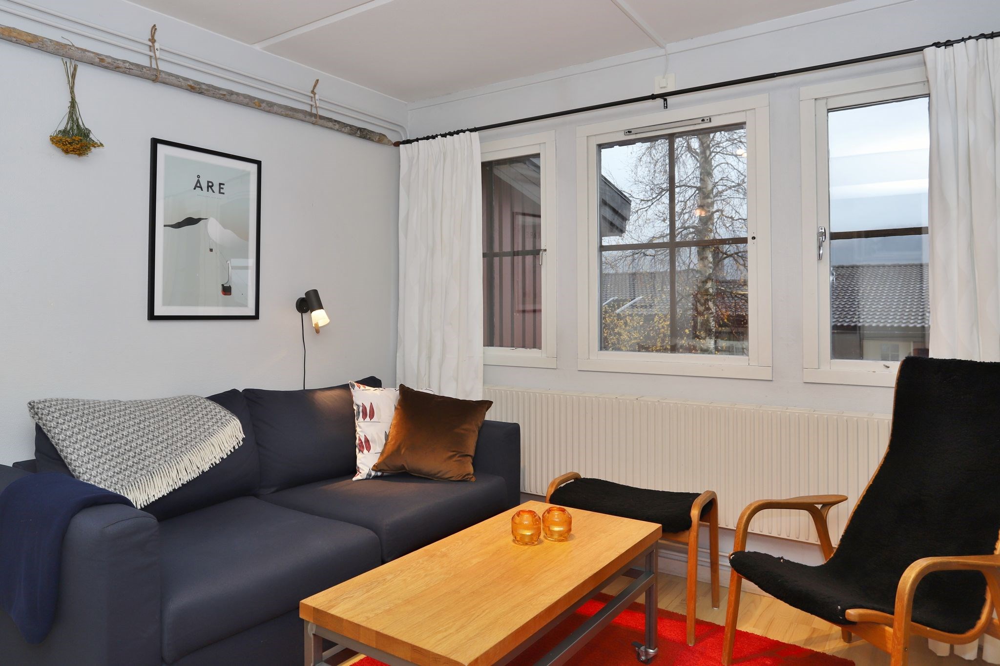 En hyggelig stue med en mørkeblå sofa, en sort lænestol, et sofabord i træ med to orange glaslamper, hvide gardiner, en væglampe og et indrammet kunstværk. Store vinduer afslører træer og hustage udenfor.