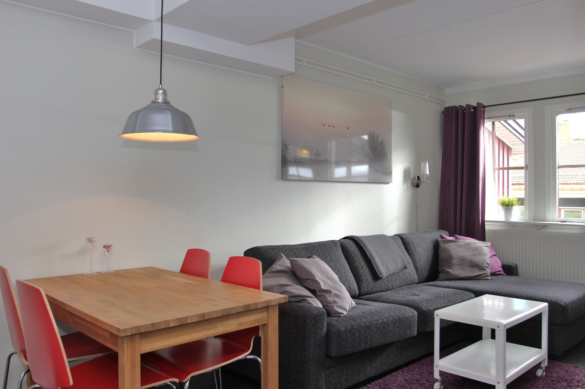 En moderne stue med en grå sofa, lilla puder, et lille hvidt sofabord og et spisebord i træ med fire røde stole. En hængelampe er over bordet, og sollyset kommer ind gennem et vindue med lilla gardiner.
