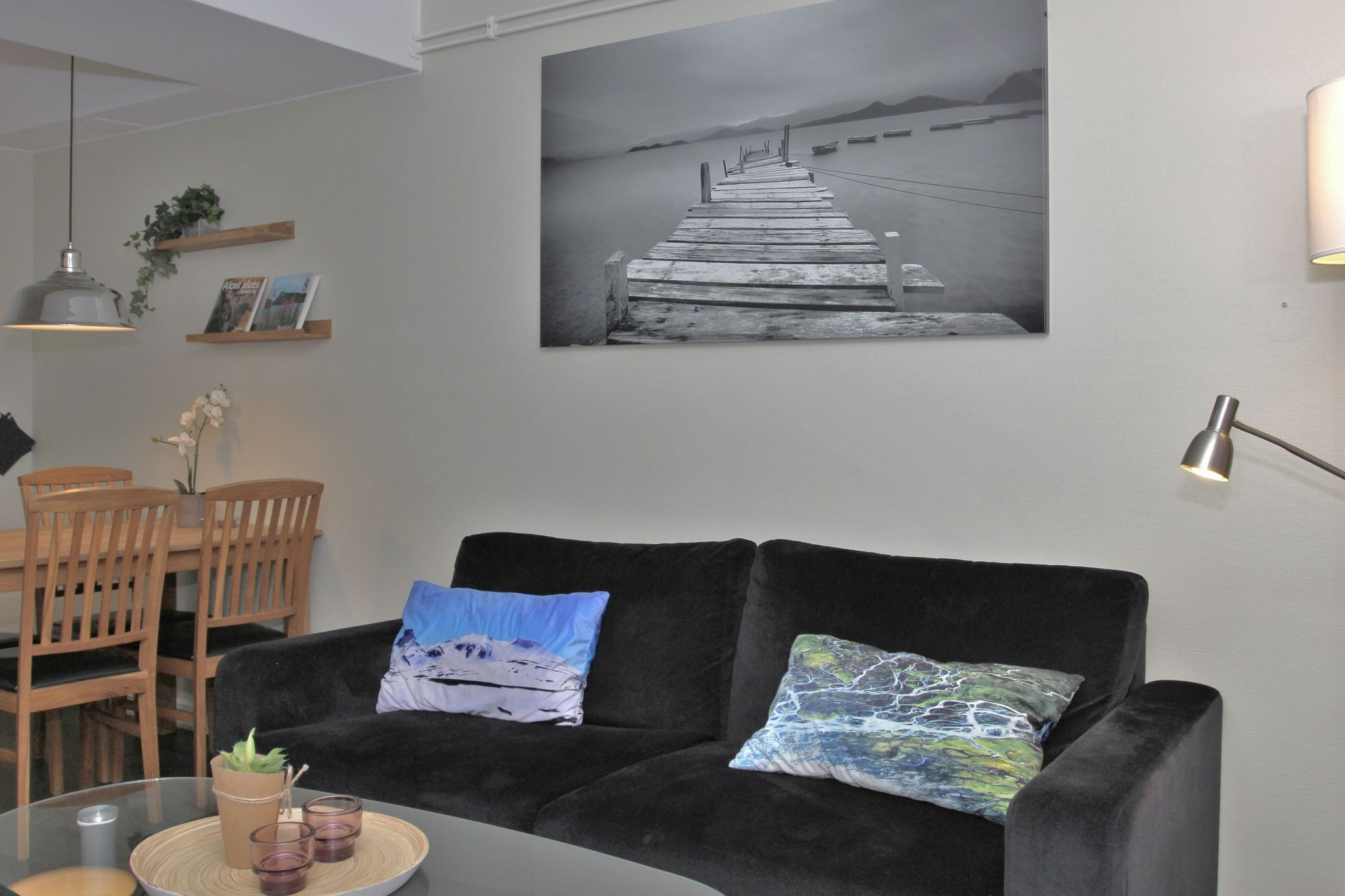 En moderne stue med en sort sofa, farverige puder, et sofabord i glas og vægkunst med et sort-hvidt foto af en træmole over vandet. Spisebord med stole og planter synlige i baggrunden.