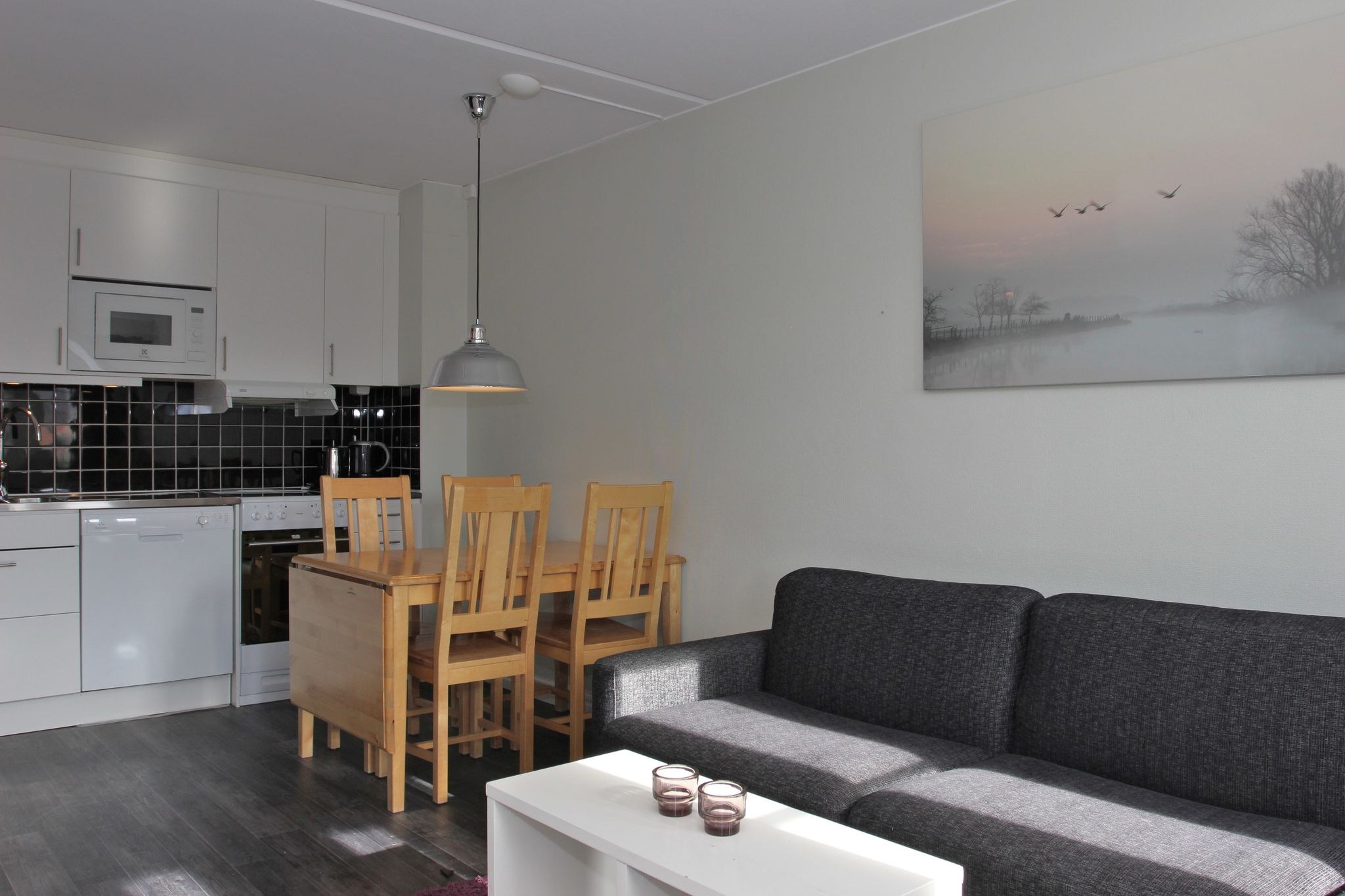En moderne lejlighedsstue med en mørkegrå sofa, et hvidt sofabord, et spisebord i træ med fire stole og et køkken med hvide skabe og mørk flisebelagt bagplade. En vægkunst og en pendellampe er også synlige.