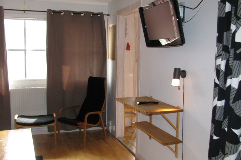 Et lille, moderne værelse med en træstol, sorte hynder, et vægmonteret skrivebord med lampe og notesblok, gardiner for et vindue og et fladskærms-tv, der står skråt fra væggen.