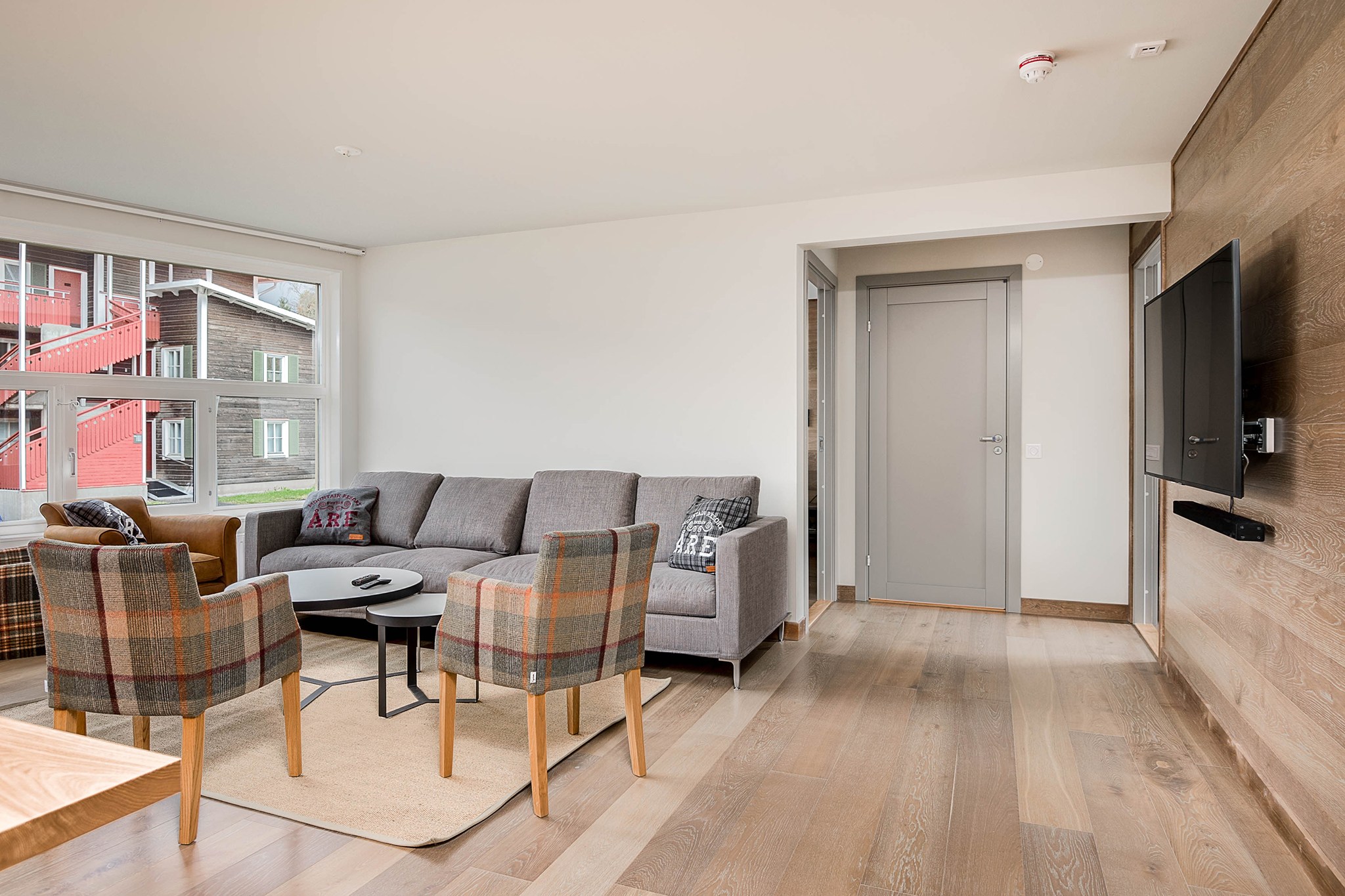 Moderne stue med lyst trægulv, grå sofa, to ternede lænestole, et rundt sofabord, vægmonteret TV og et stort vindue, der lukker naturligt lys ind. Lyse vægge og minimalistisk indretning skaber en hyggelig og luftig fornemmelse.