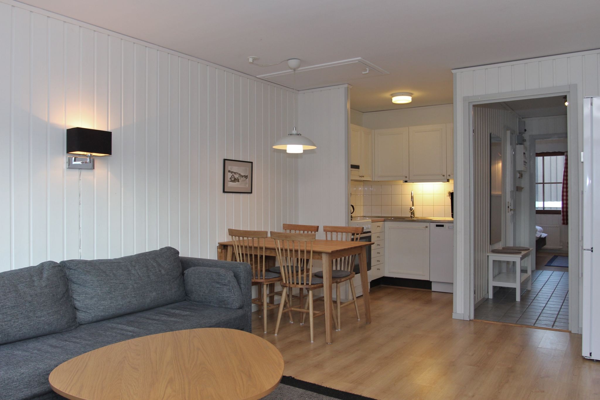 En hyggelig, moderne opholdsstue med en grå sofa, et rundt sofabord i træ, et spisebord med stole, hvide paneler på væggene og et åbent køkkenområde med hvide skabe og flisebelagt gulv.