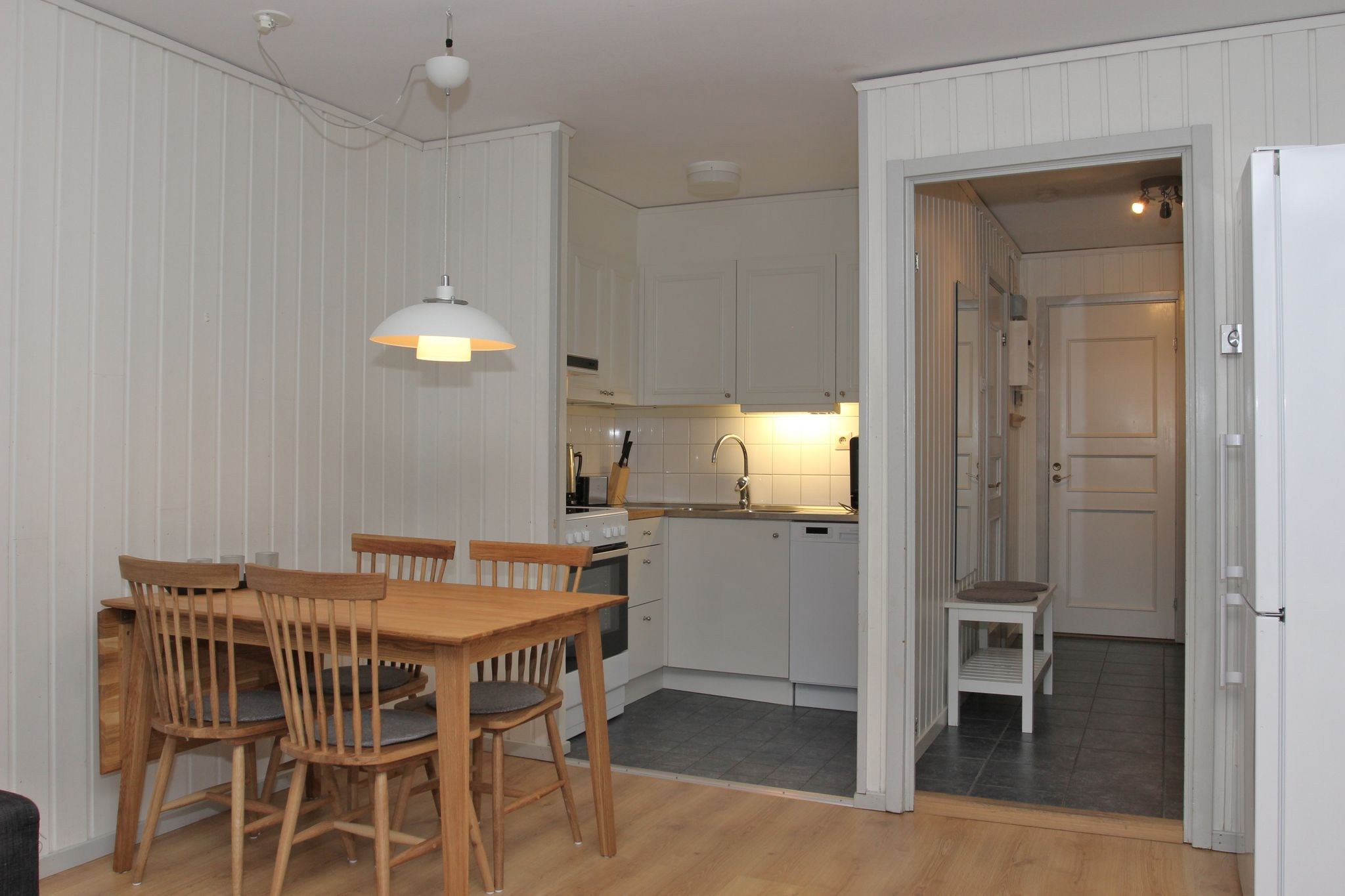 Et lille, minimalistisk køkken og spisestue med lyse træpaneler, et træbord med fire stole, hvide skabe og moderne apparater. En gang med døre er synlig gennem en åben døråbning.