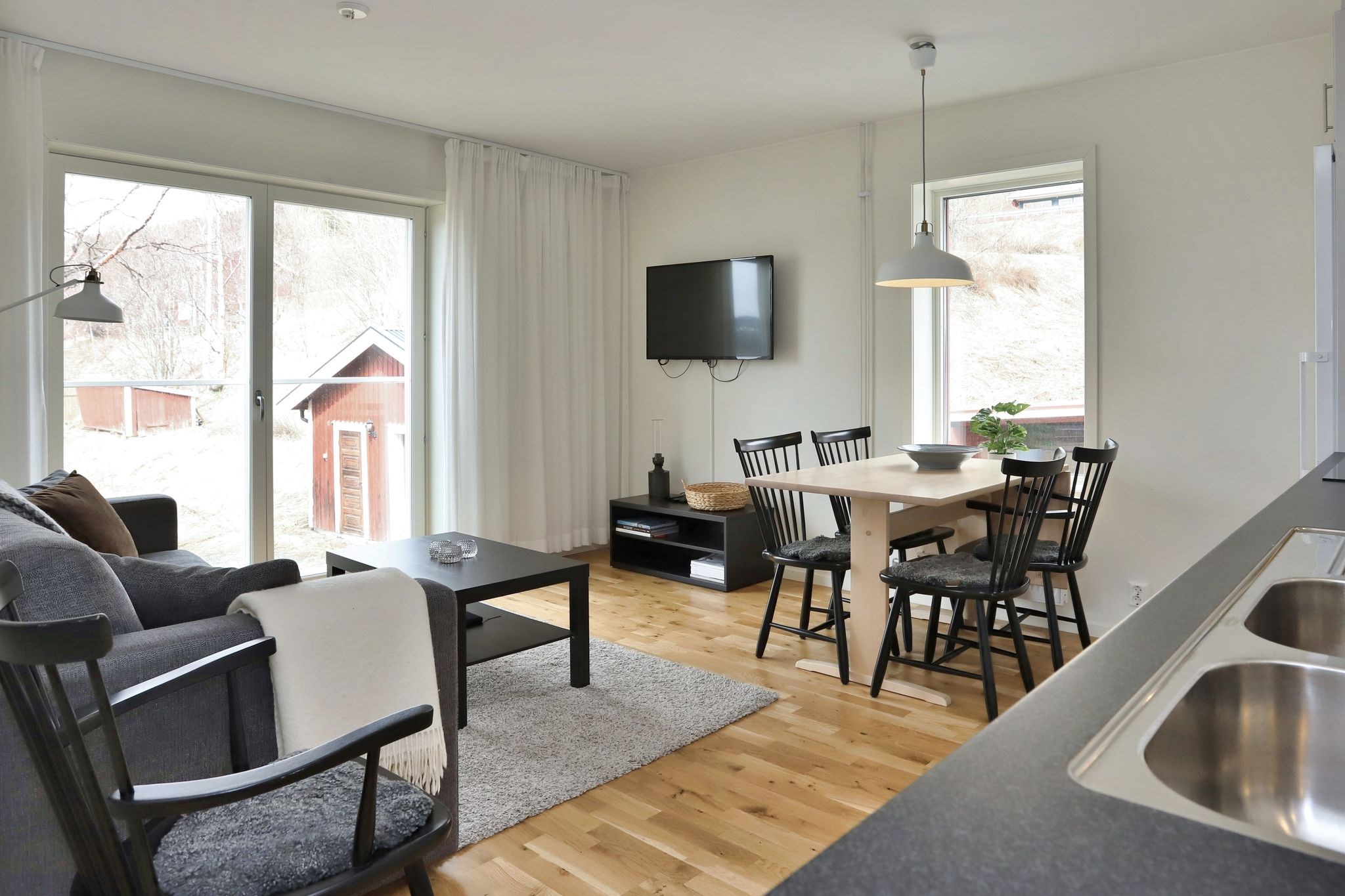 Lys, moderne opholds- og spisestue med trægulv, grå sofa, lille spisebord med fire sorte stole, vægmonteret TV, store vinduer og lys, der strømmer ind udefra.