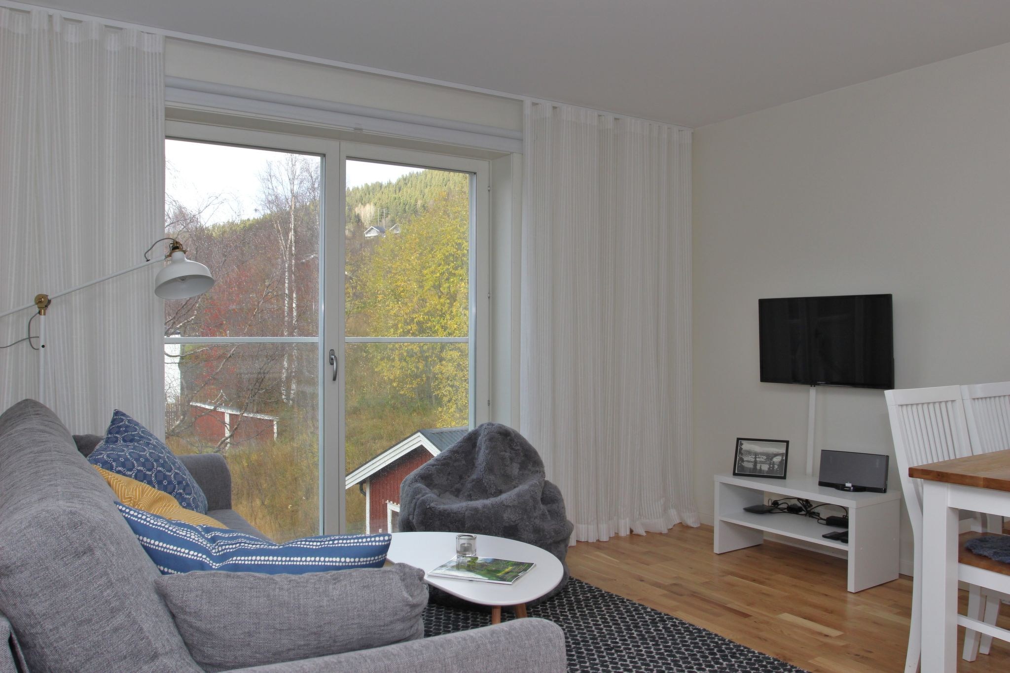En hyggelig stue med en grå sofa, mønstrede puder, et lille rundt bord, en gulvlampe, en sækkestol, et tv på et hvidt bord og store vinduer med tynde gardiner med udsigt over træer og en rød bygning udenfor.