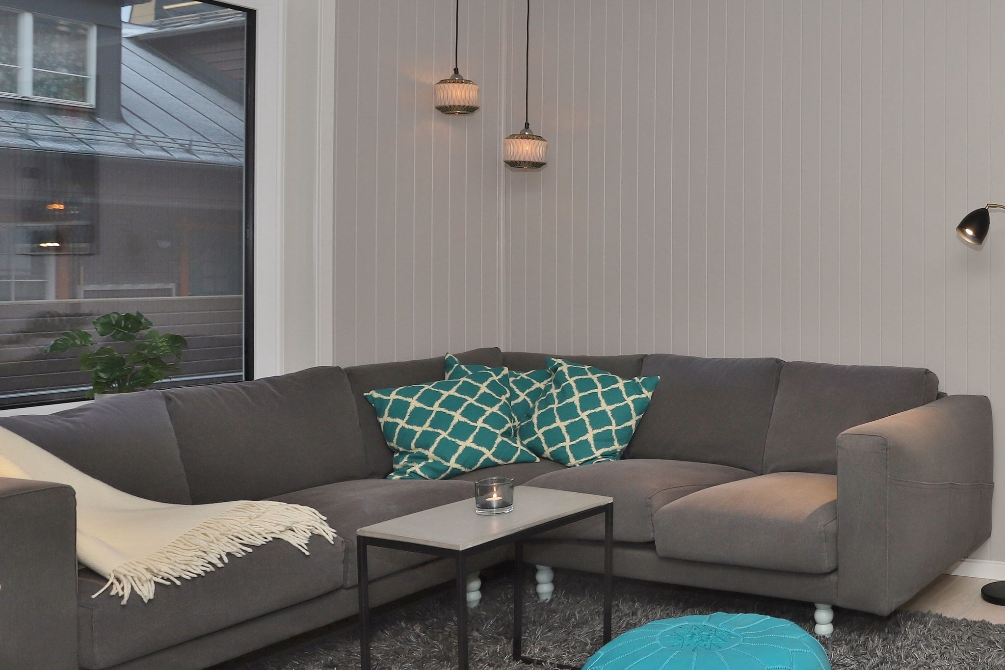 En moderne stue med en grå sofa, hynder med mønstre i blågrøn og hvidt, et hvidt tæppe, et lille sofabord med et stearinlys, en blå puf, pendellamper og et stort vindue.