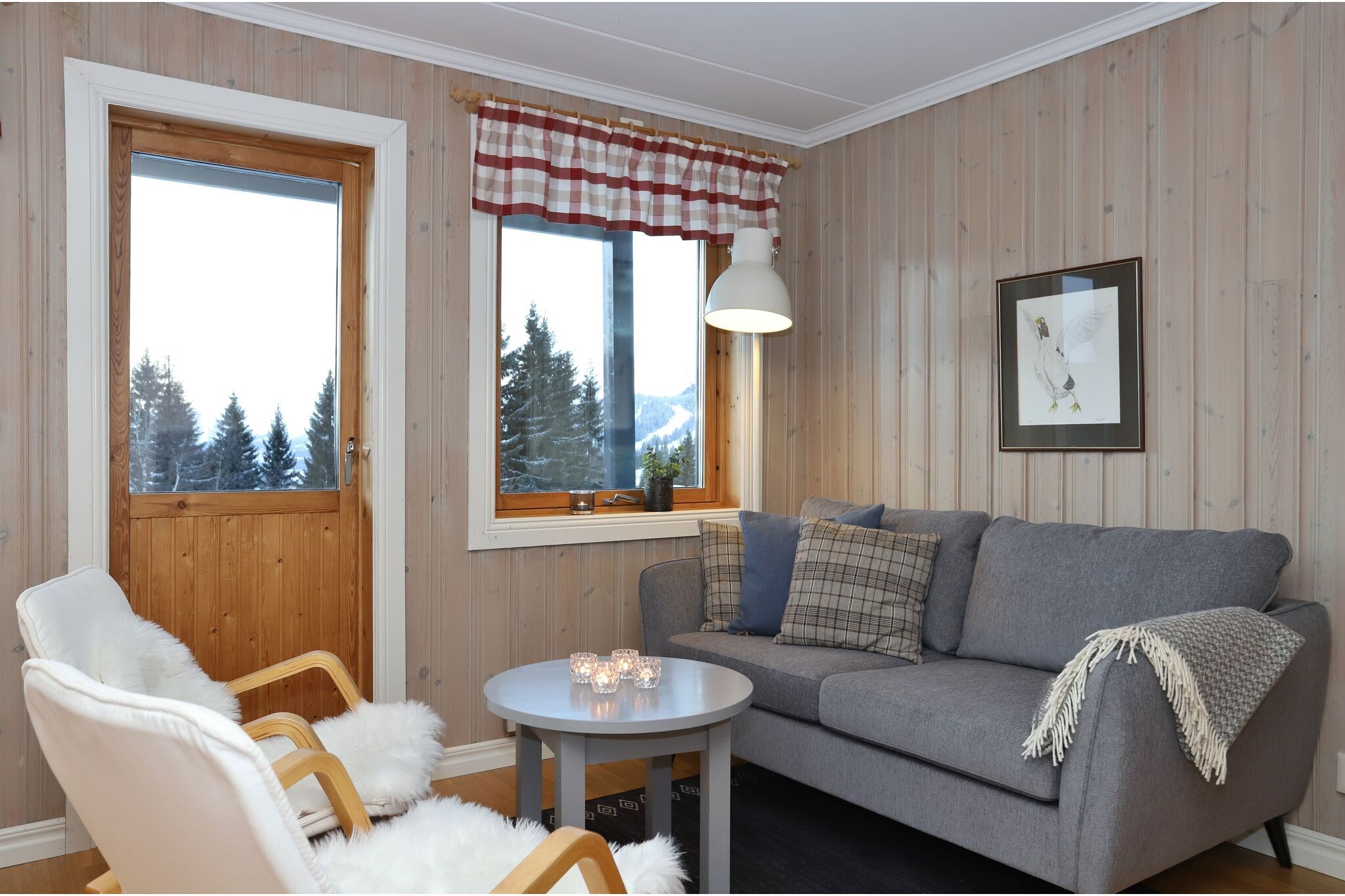 En hyggelig stue med trævægge, en grå sofa, to hvide lænestole, et rundt sofabord med stearinlys, et indrammet billede på væggen og et vindue med et rødt ternet gardin, der viser en snedækket bjergudsigt.