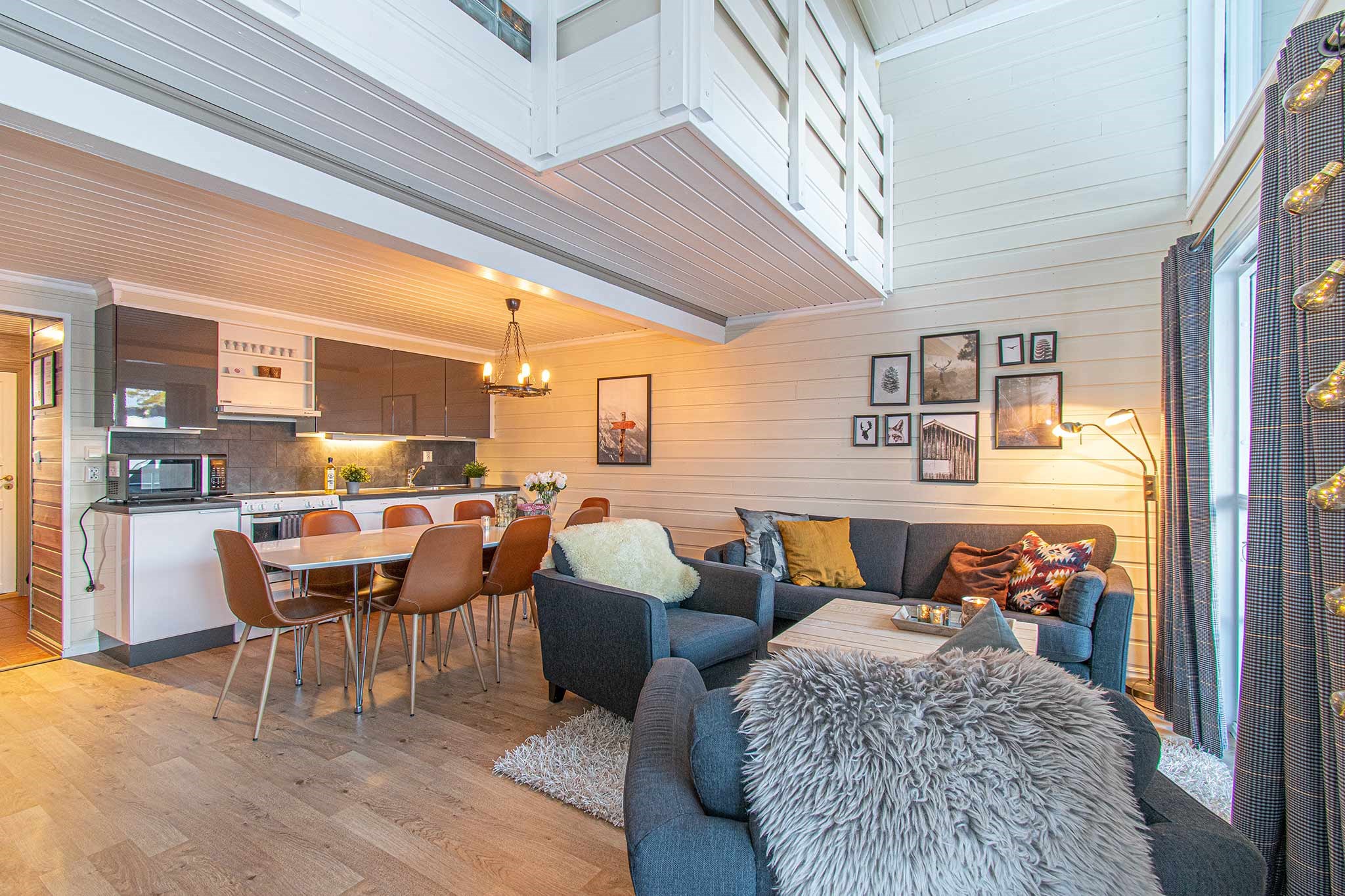 En hyggelig, moderne åben stue og køkken med trævægge, et spisebord med seks stole, en grå sofa og lænestole, vægkunst, et køkkenområde med hvide skabe og varm belysning.