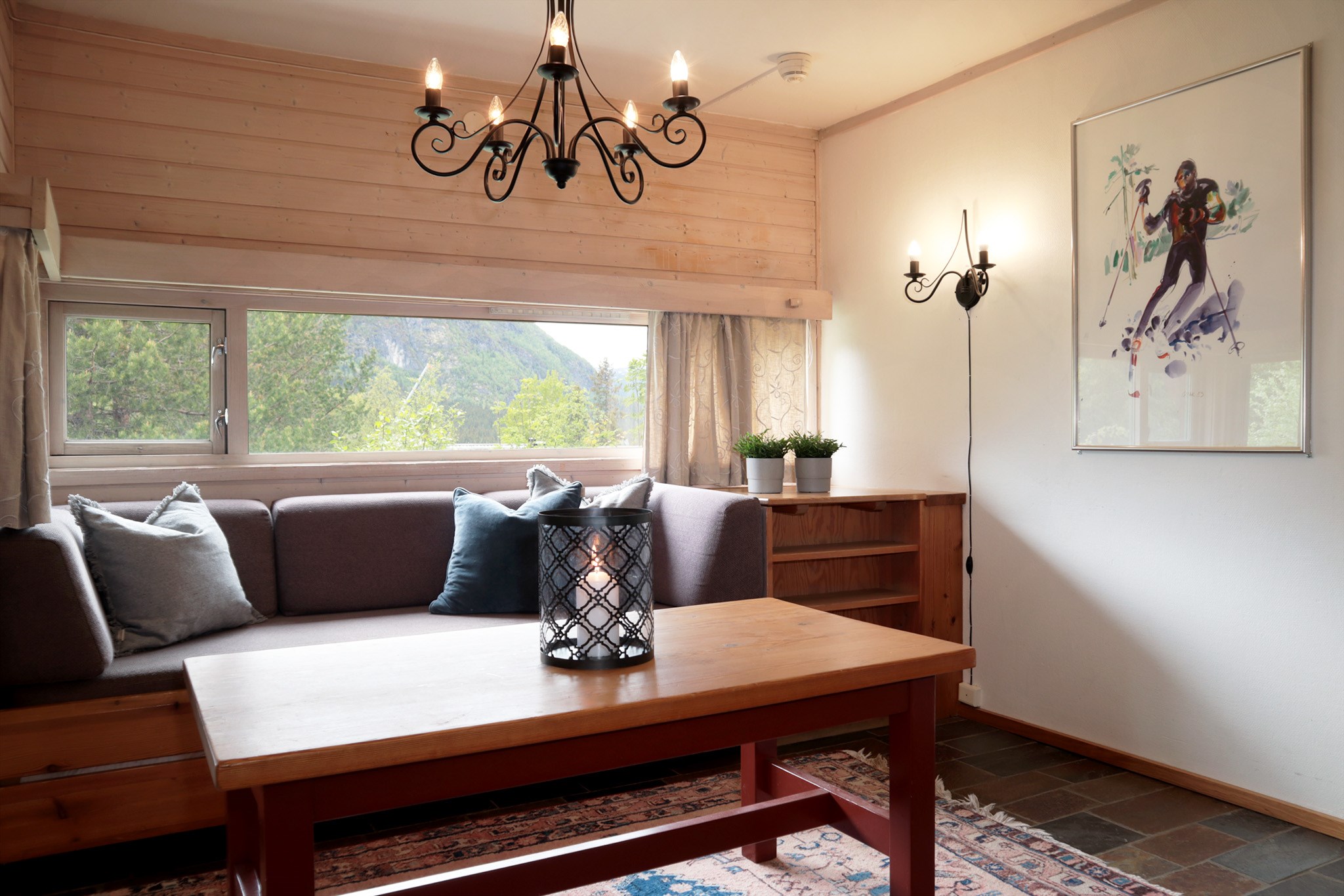Hyggelig stue med et træbord, grå sofa med puder, stort vindue med bjergudsigt, vægkunst med en skiløber, midtpunktsdekoration som stearinlys, potteplanter og sorte metallamper.