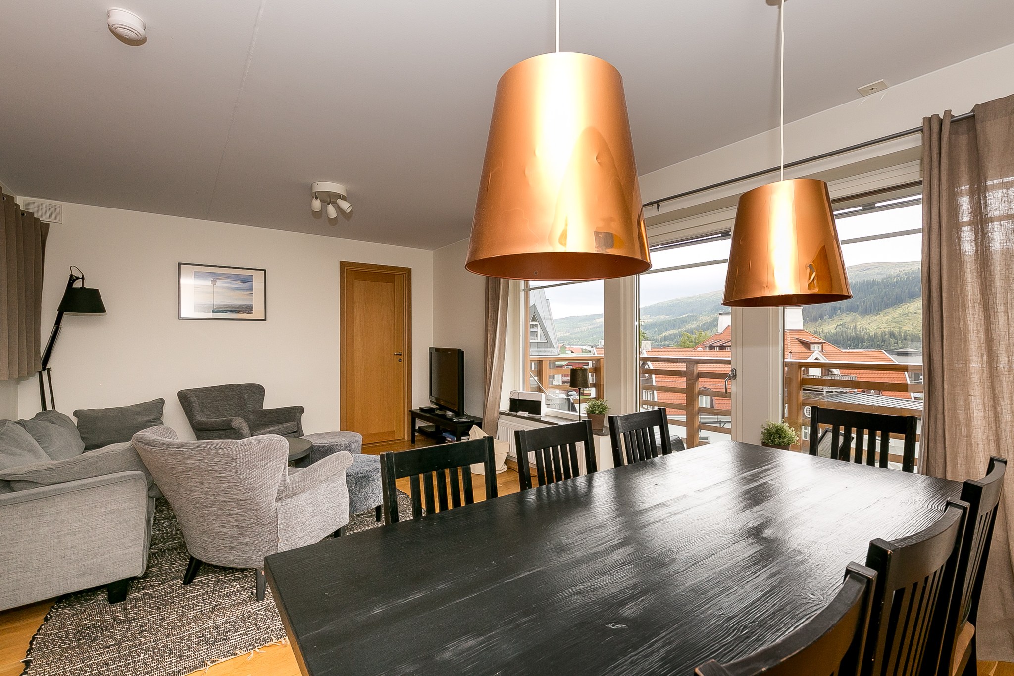En moderne opholds- og spisestue med et sort bord, mørke stole, grå sofaer, store kobberpendler og vinduer med en smuk udsigt over bjergene.