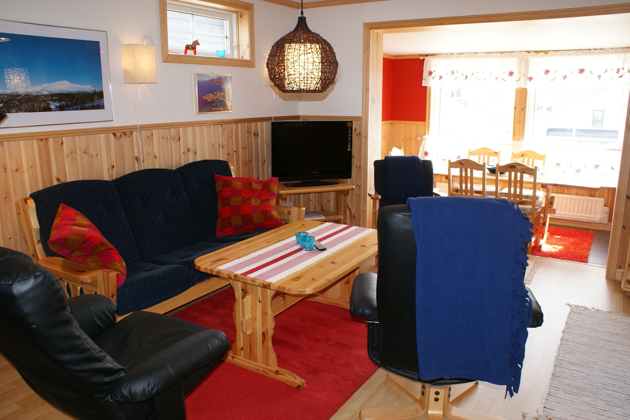 Hyggelig stue med træpaneler på væggene, marineblå sofa, to sorte stole, sofabord i træ, TV og lys spiseplads i baggrunden ved store vinduer med naturligt lys. Røde accenter overalt.