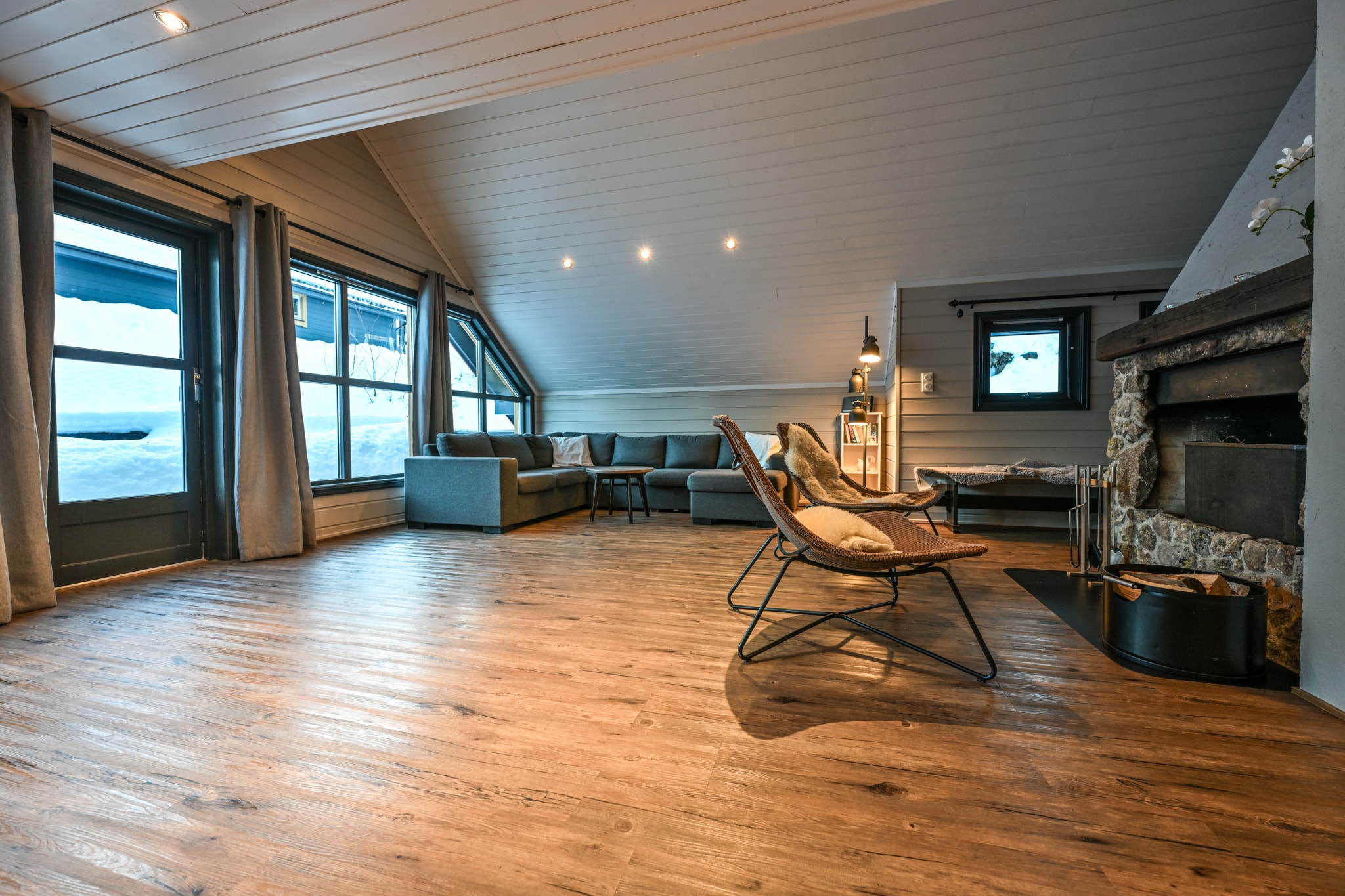 En hyggelig stue med trægulve, moderne møbler, en stenpejs, store vinduer med gardiner og et skrånende loft. Sneen kan ses udenfor gennem vinduerne.