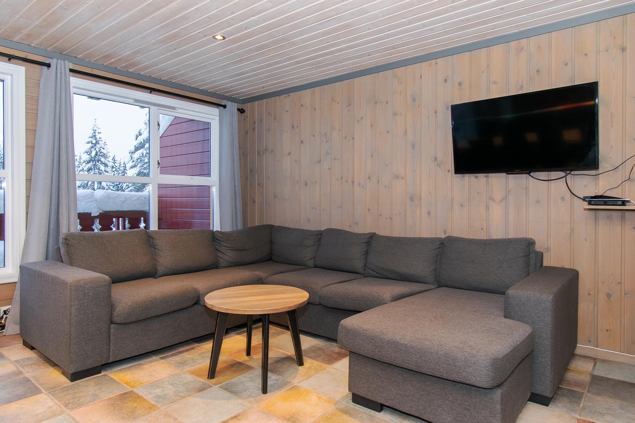 En hyggelig stue med en stor L-formet grå sofa, et rundt sofabord i træ, et vægmonteret tv og træpaneler på væggene; et vindue viser snedækkede træer og en rød balkon udenfor.