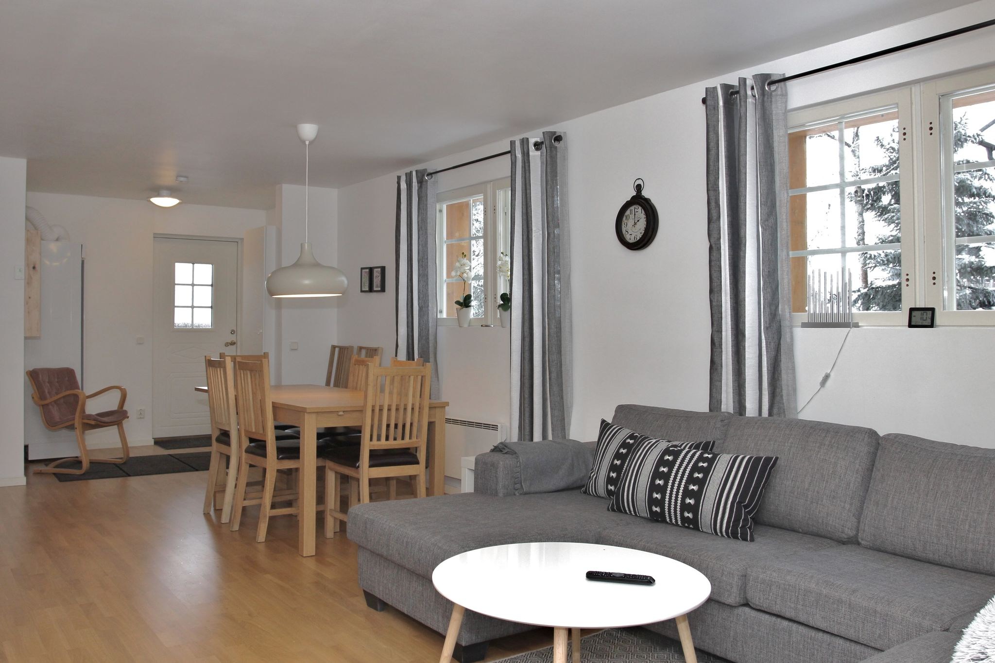 En moderne opholds- og spisestue med lyse trægulve, en grå sofa med mønstrede puder, et rundt hvidt sofabord, et spisebord med seks stole, grå gardiner og store vinduer, der lukker naturligt lys ind.