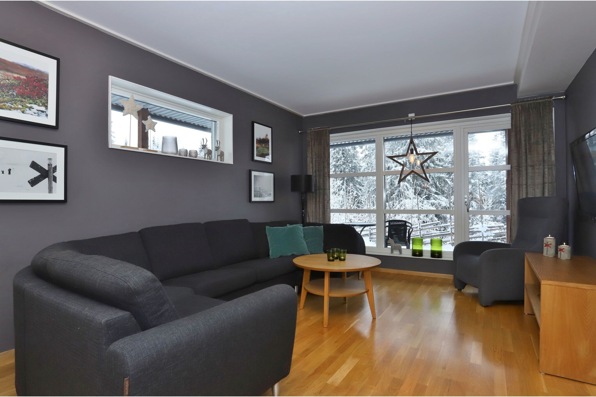 En moderne stue med mørkegrå vægge, en sort sofa, et rundt sofabord i træ og store vinduer, der viser et snedækket udendørsscene. Dekorationerne omfatter indrammet kunst, grønne puder og en stjerneformet lampe.