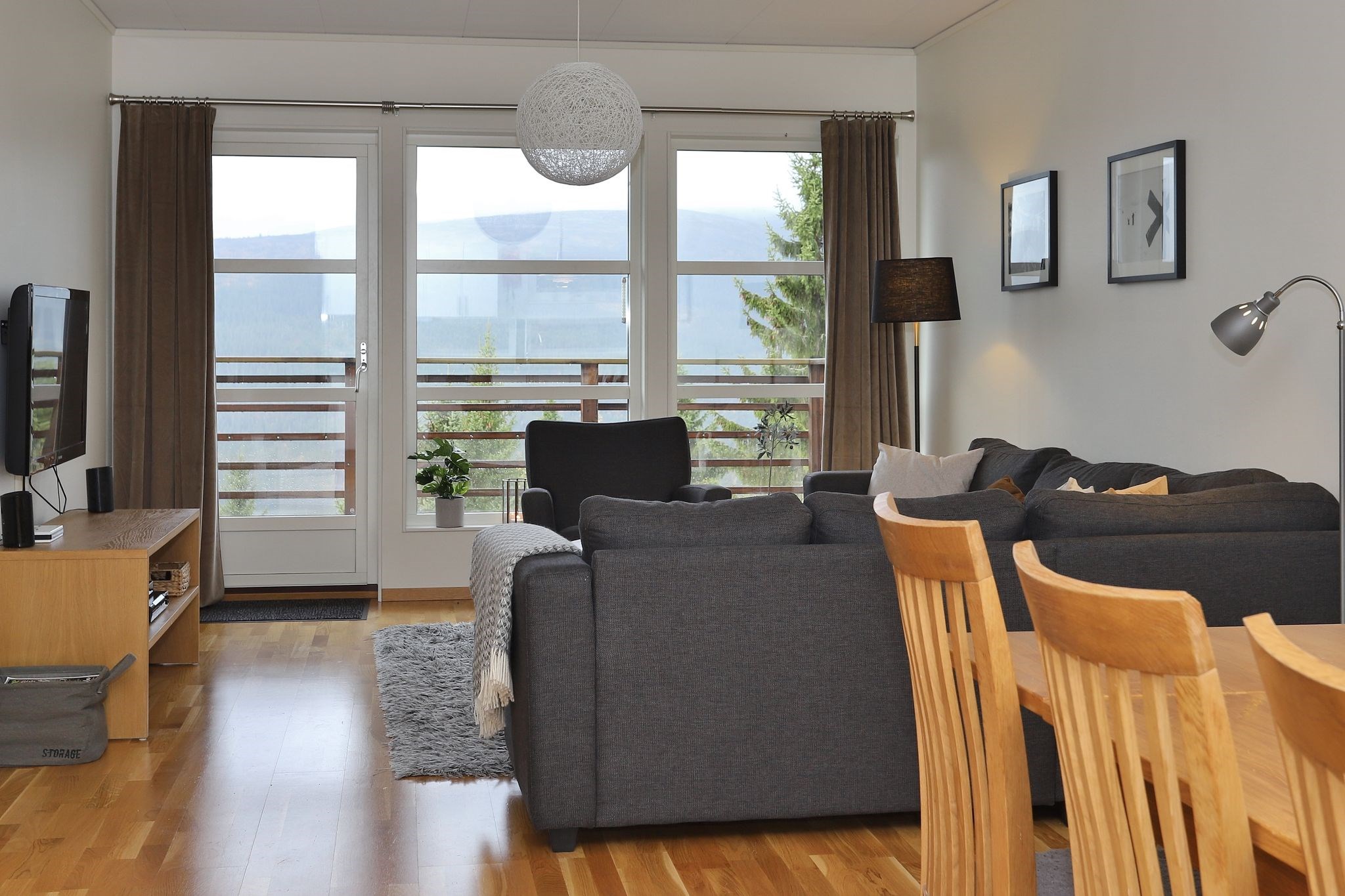 En moderne stue med en grå hjørnesofa, et spisebord i træ med stole, TV på et stativ, gulvlampe og store vinduer, der fører ud til en balkon med en smuk udsigt.