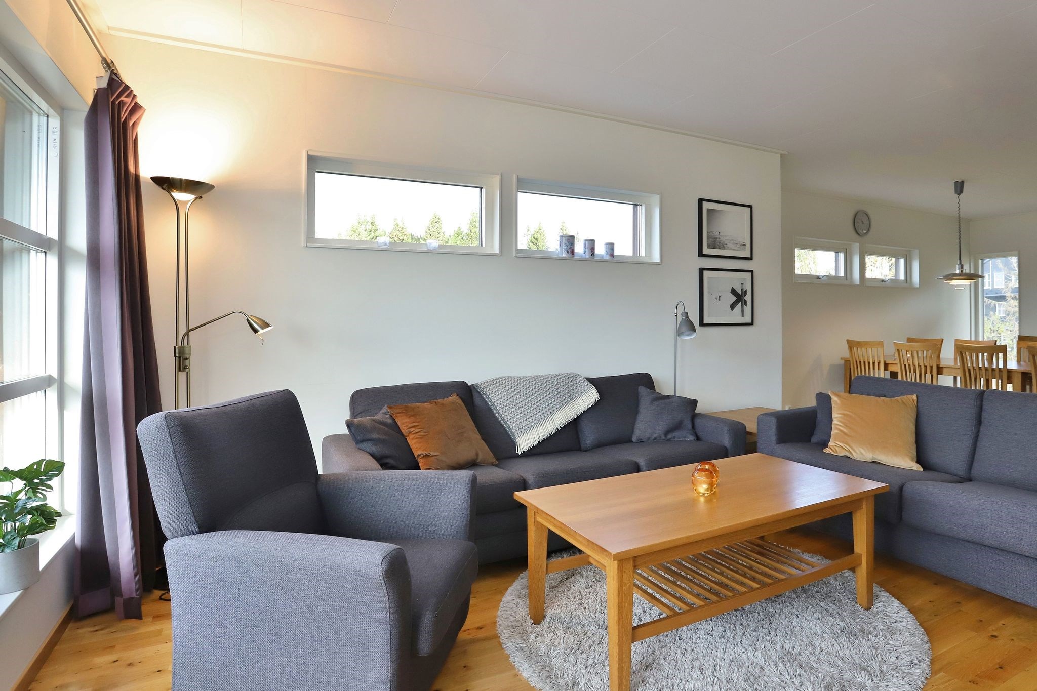 En moderne stue med grå sofaer, et sofabord i træ, en grå lænestol, en gulvlampe, vægkunst og et spisebord i baggrunden. Store vinduer lukker naturligt lys ind.