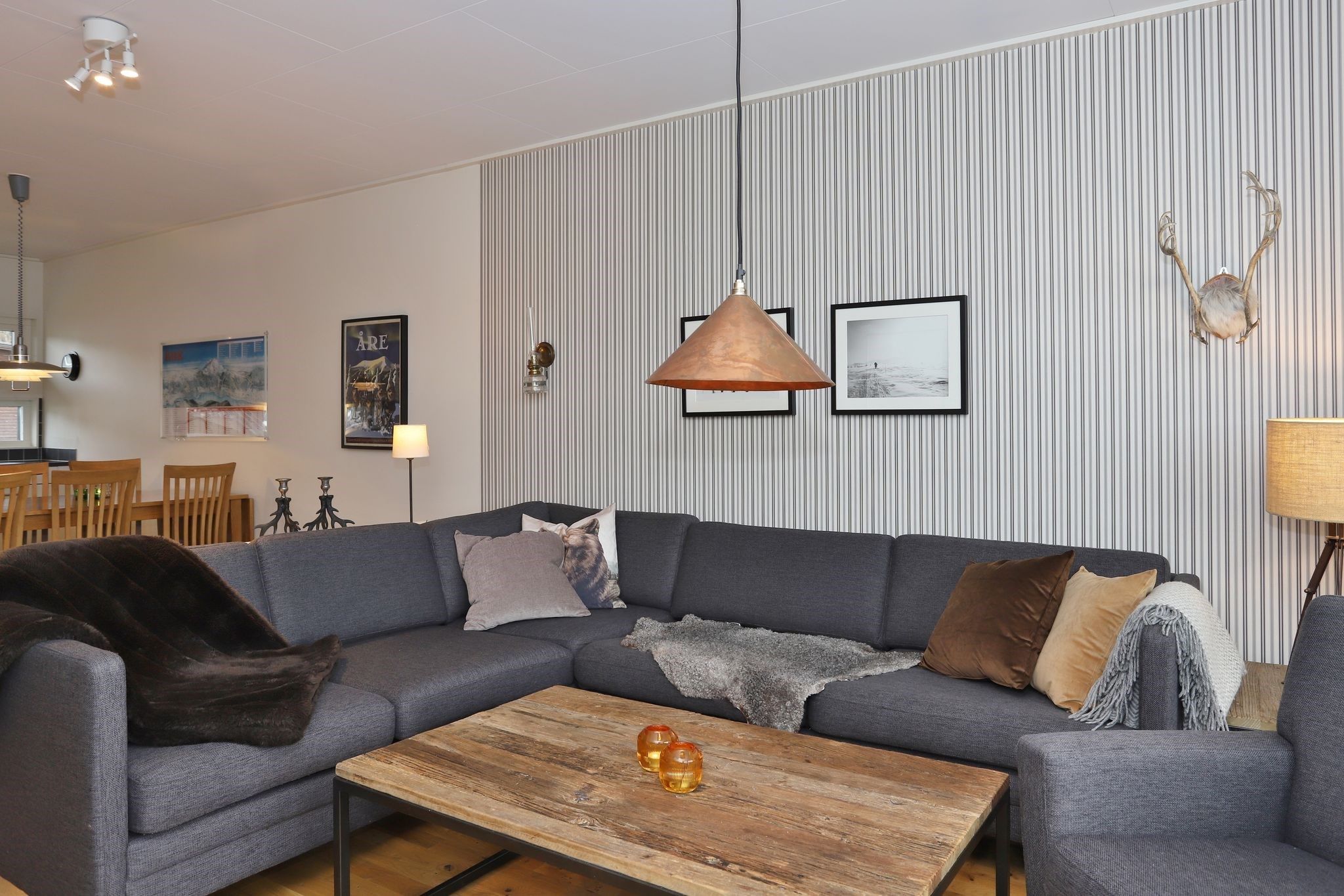 En moderne stue med en grå sofa, et sofabord i træ og to små orange stearinlys. Værelset har indrammet kunst, en pendellampe, vægdekoration med gevir og en spiseplads i baggrunden.