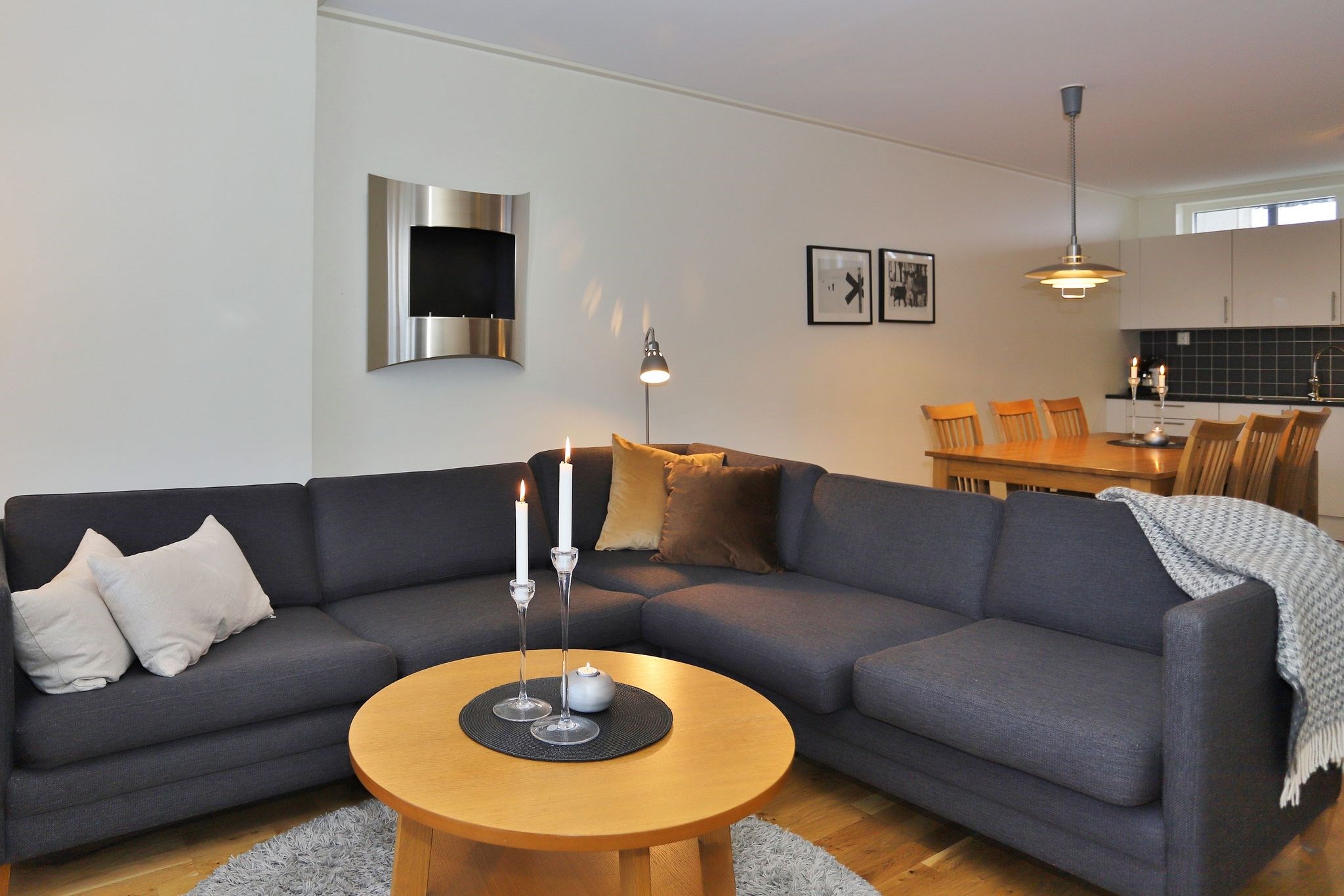 Moderne stue med en mørkegrå sofa, lyse og mørke puder, et rundt sofabord i træ med stearinlys og en spiseplads i baggrunden. Neutrale farver og enkel, hyggelig indretning.