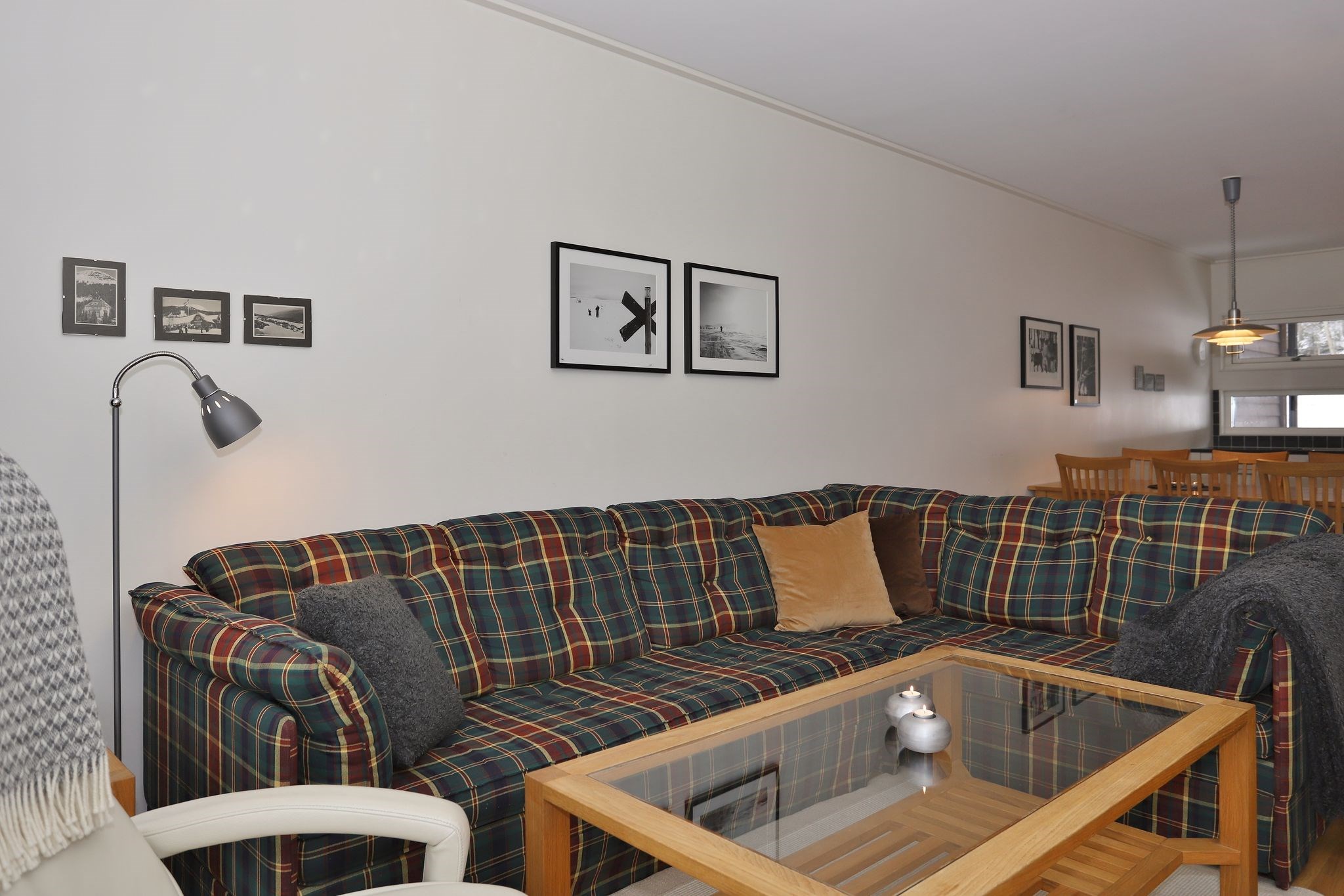 En stue med en ternet hjørnesofa, et sofabord i glas, vægkunst med sort-hvide fotos, en gulvlampe og en spiseplads med et træbord og stole i baggrunden.